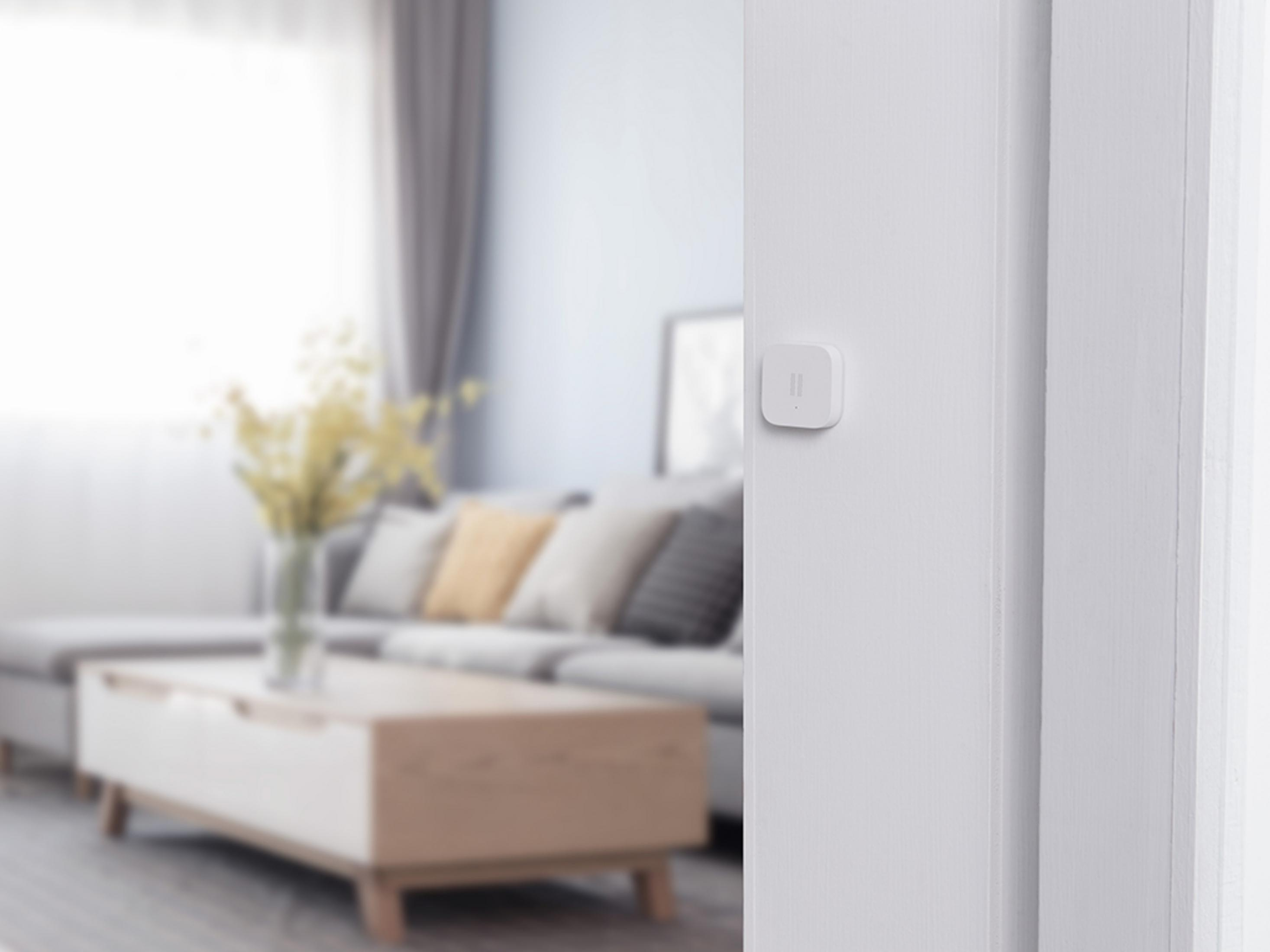 Ein kleines weißes Smart-Home-Gerät, das an einer weißen Wand montiert ist, mit einem verschwommenen Wohnzimmer im Hintergrund.