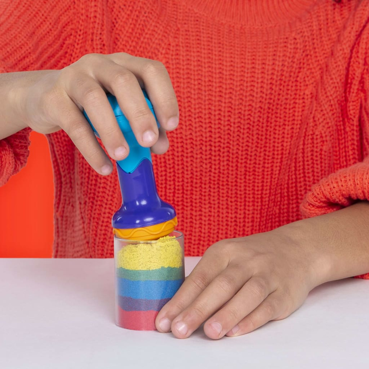 Une main utilise un outil bleu pour emballer Kinetic Sand dans un récipient.