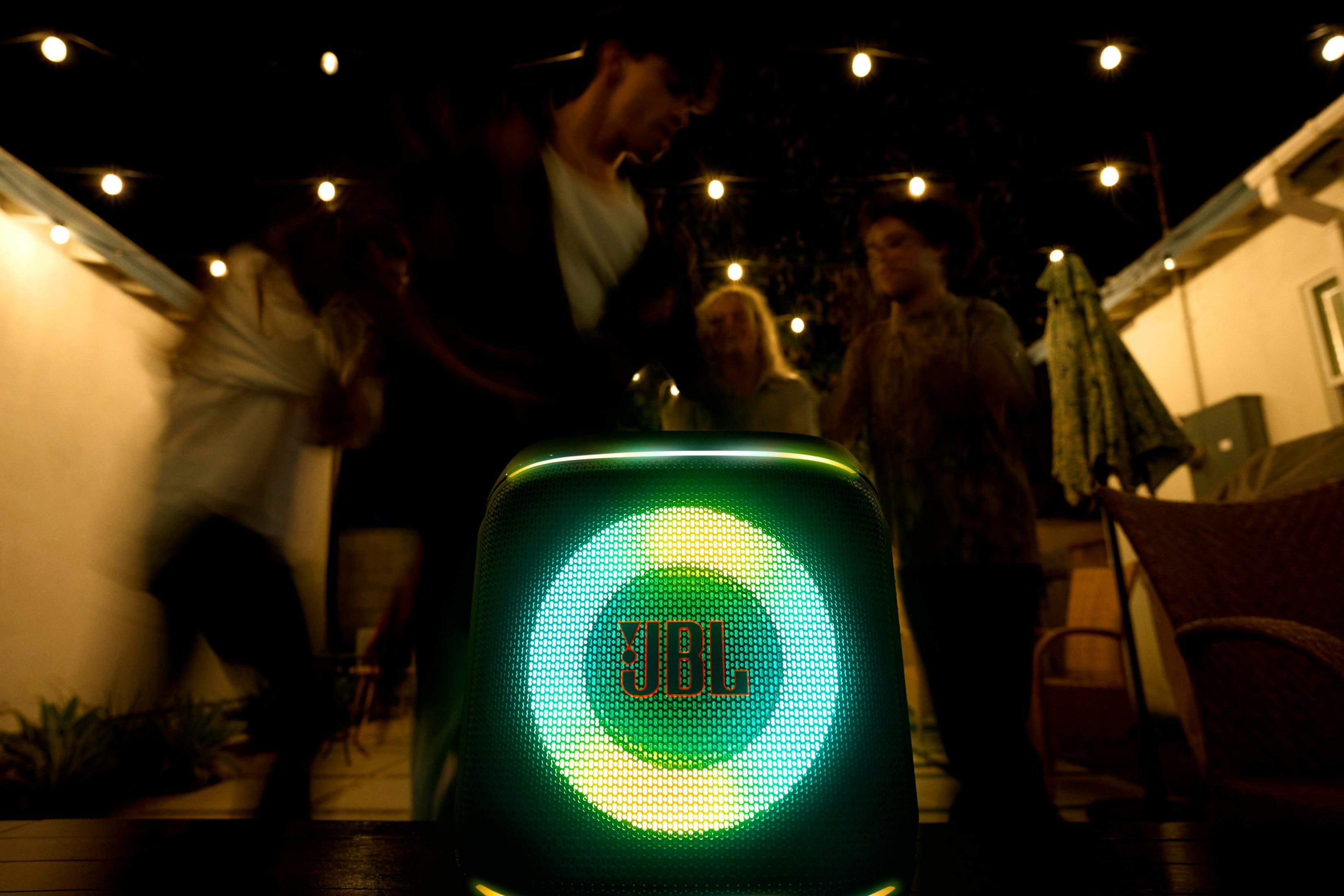 Un altoparlante JBL si illumina di luci verdi e gialle mentre le persone ballano sullo sfondo sotto le luci di una stringa di notte.