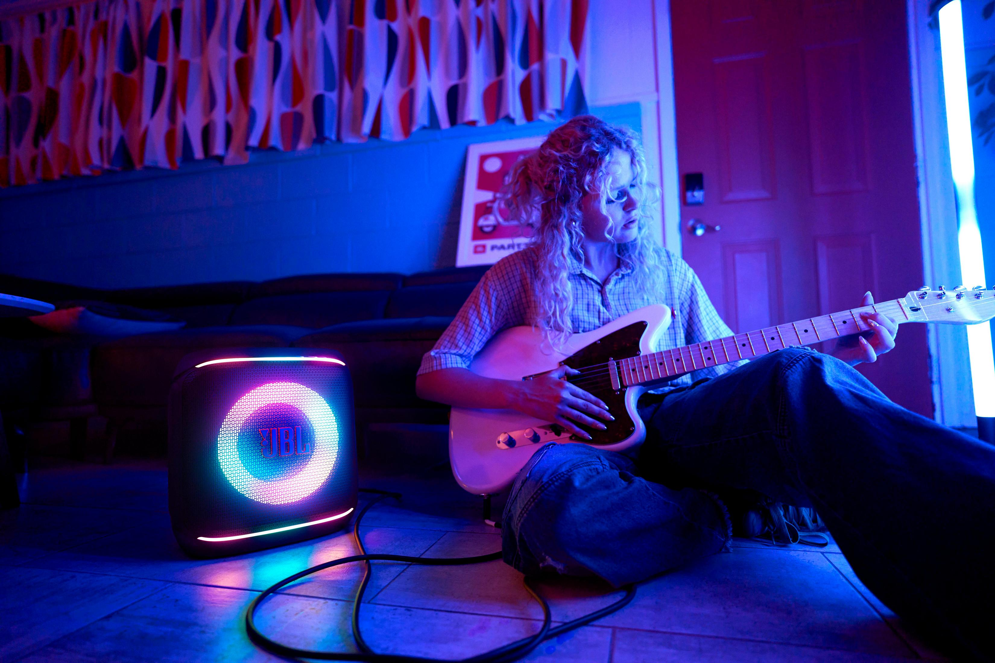 Una persona suona una chitarra elettrica bianca accanto a un altoparlante JBL con luci colorate in una stanza con illuminazione blu.