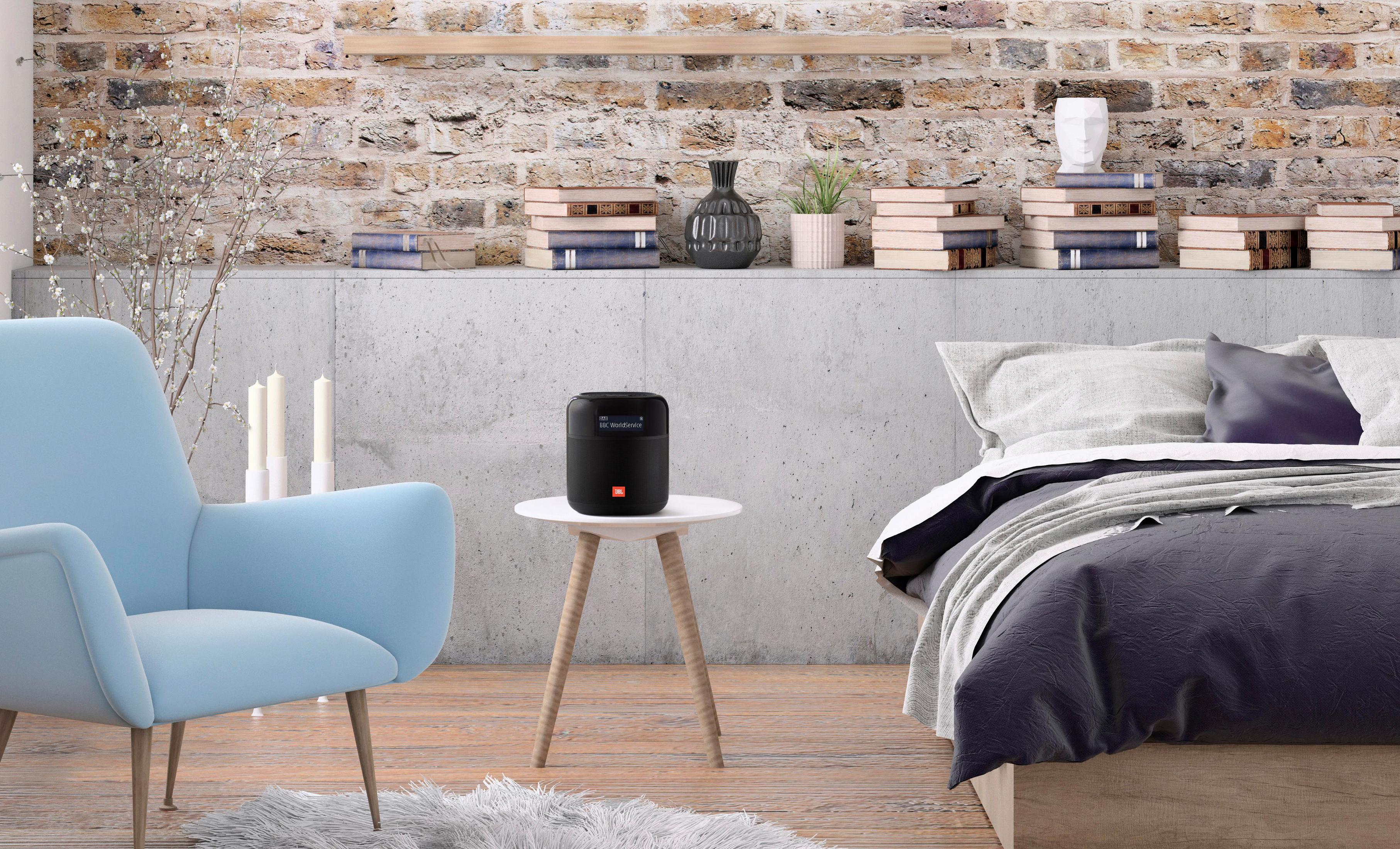 Un altoparlante JBL nero è appoggiato su un tavolo in una camera da letto accogliente con una sedia blu e un muro di mattoni.