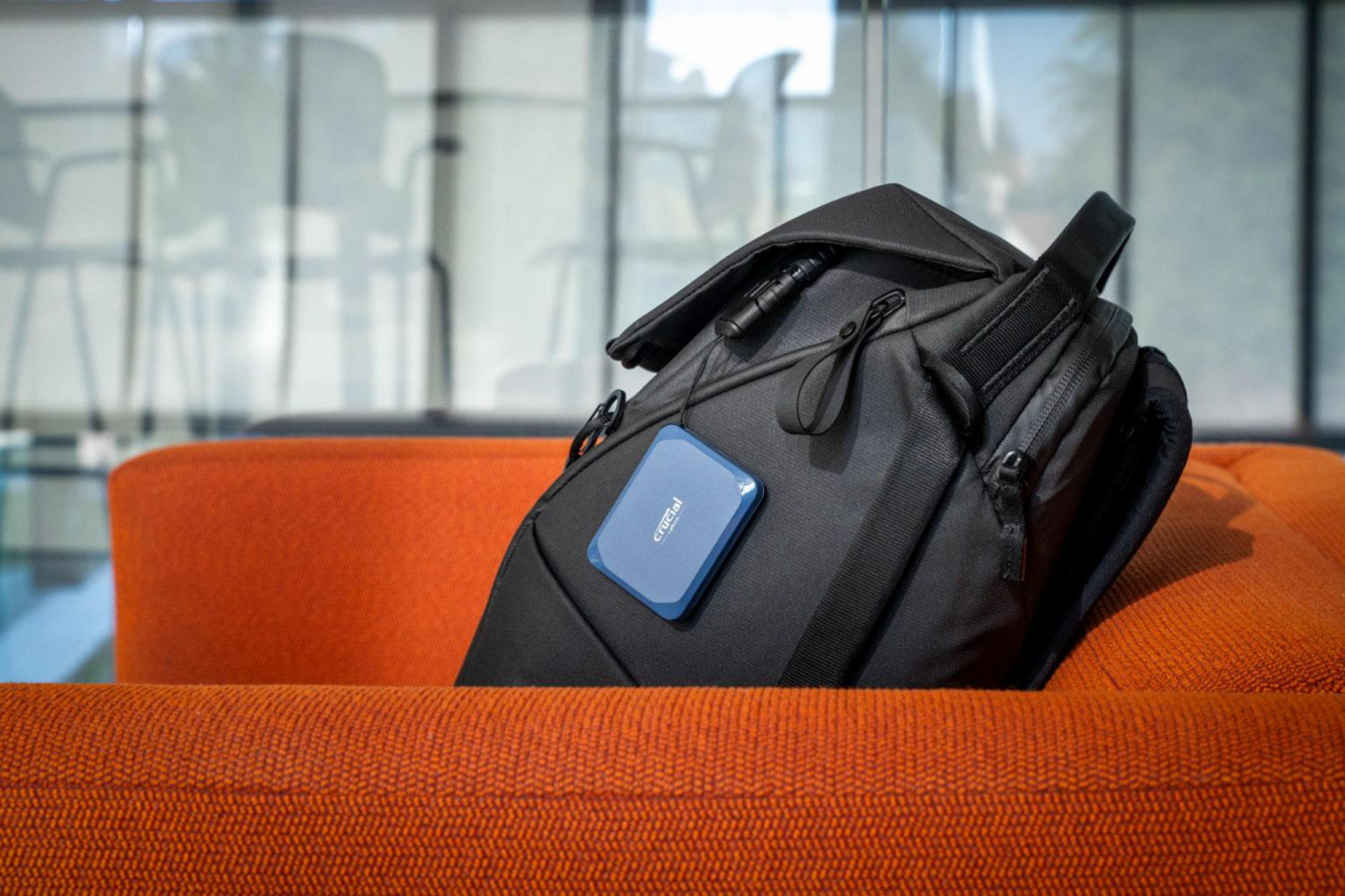 Niebieski dysk SSD Crucial wisi na czarnym plecaku na pomarańczowej kanapie na tle rozmazanego szkła.