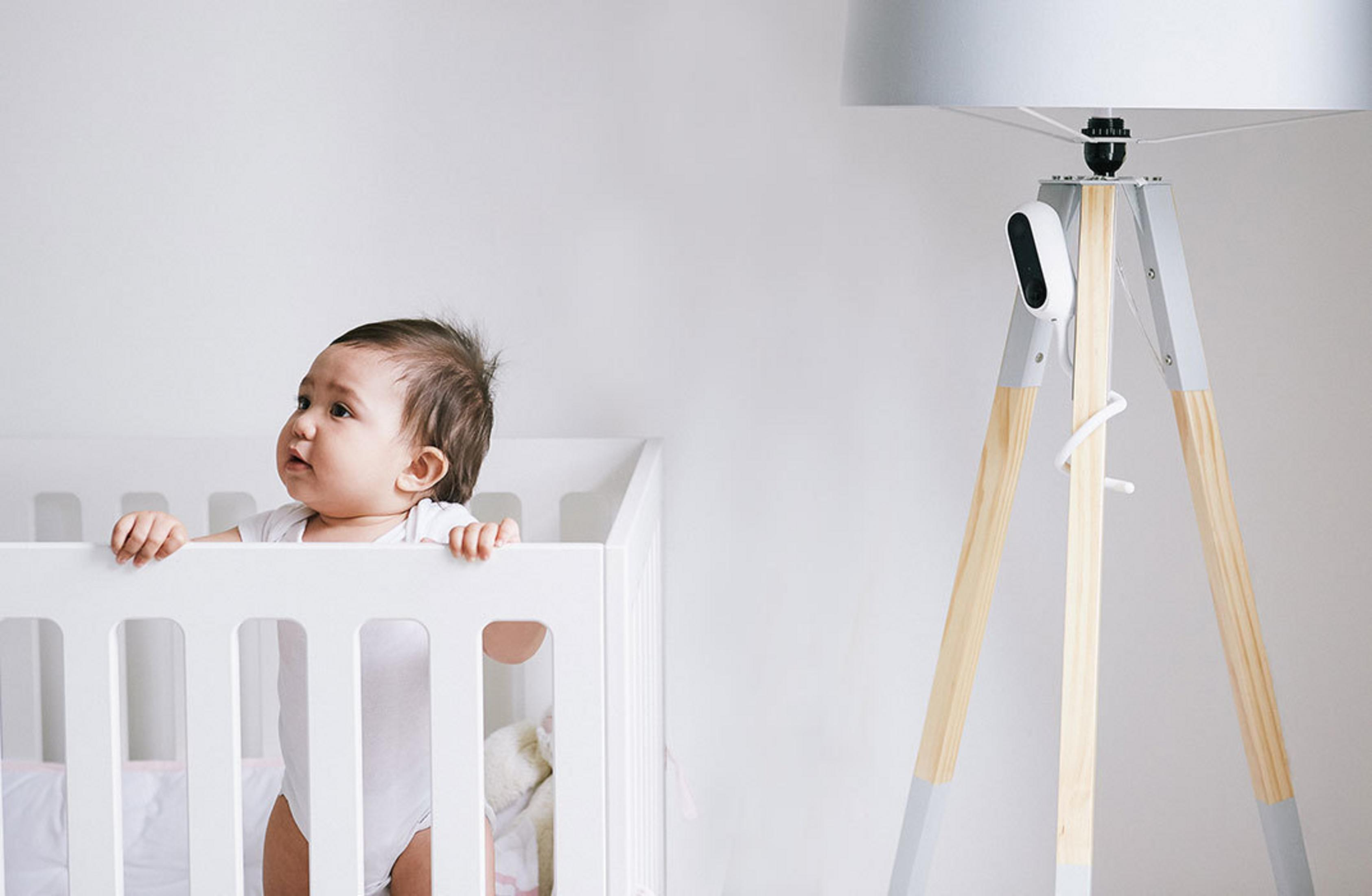 Baby in einem weißen Kinderbett, das eine Canary-Überwachungskamera ansieht, die auf einer Stehlampe vor einer weißen Wand steht.