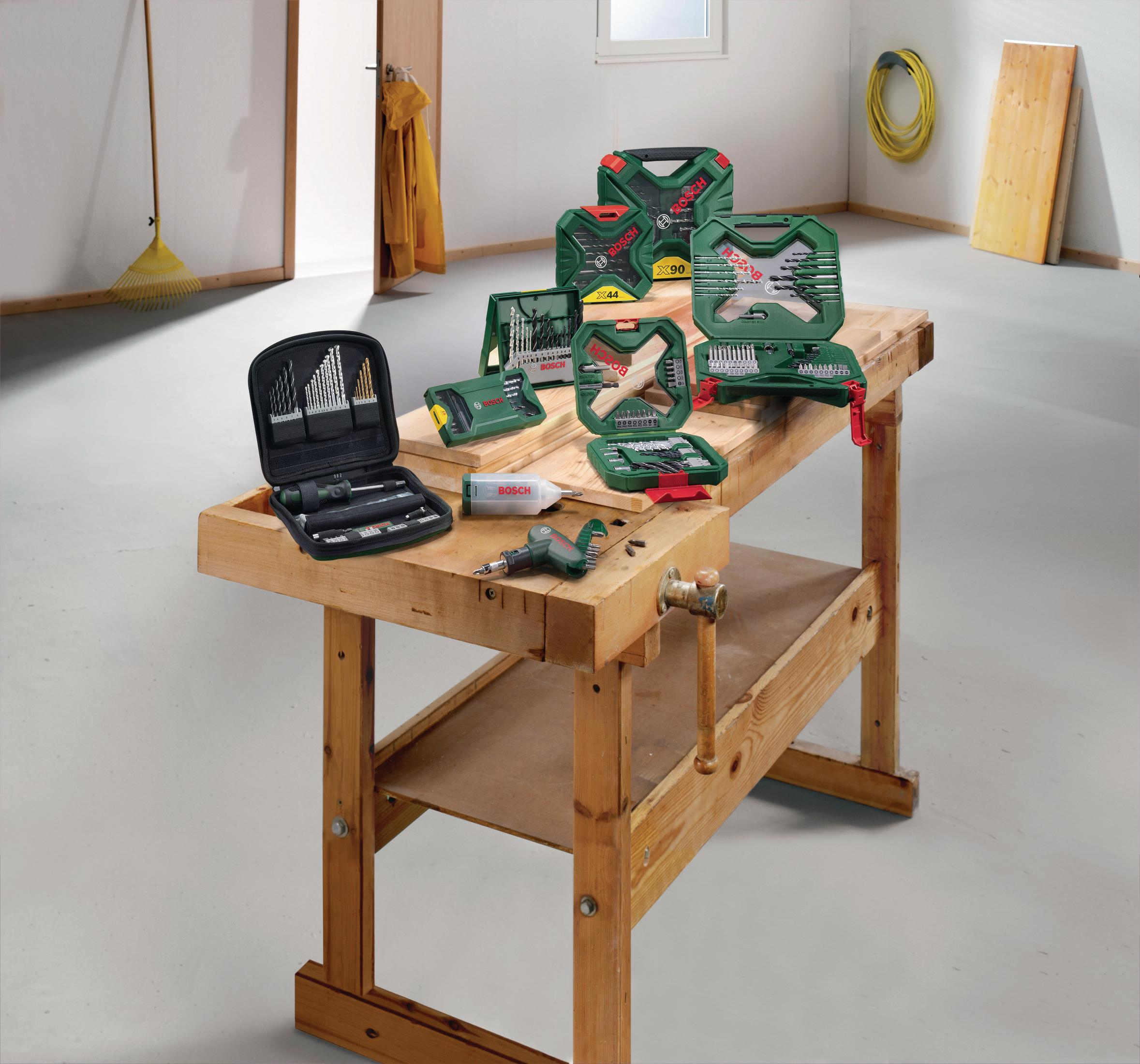 Des jeux d'outils Bosch sont disposés sur un établi en bois dans une pièce avec un mur blanc et un sol en béton.