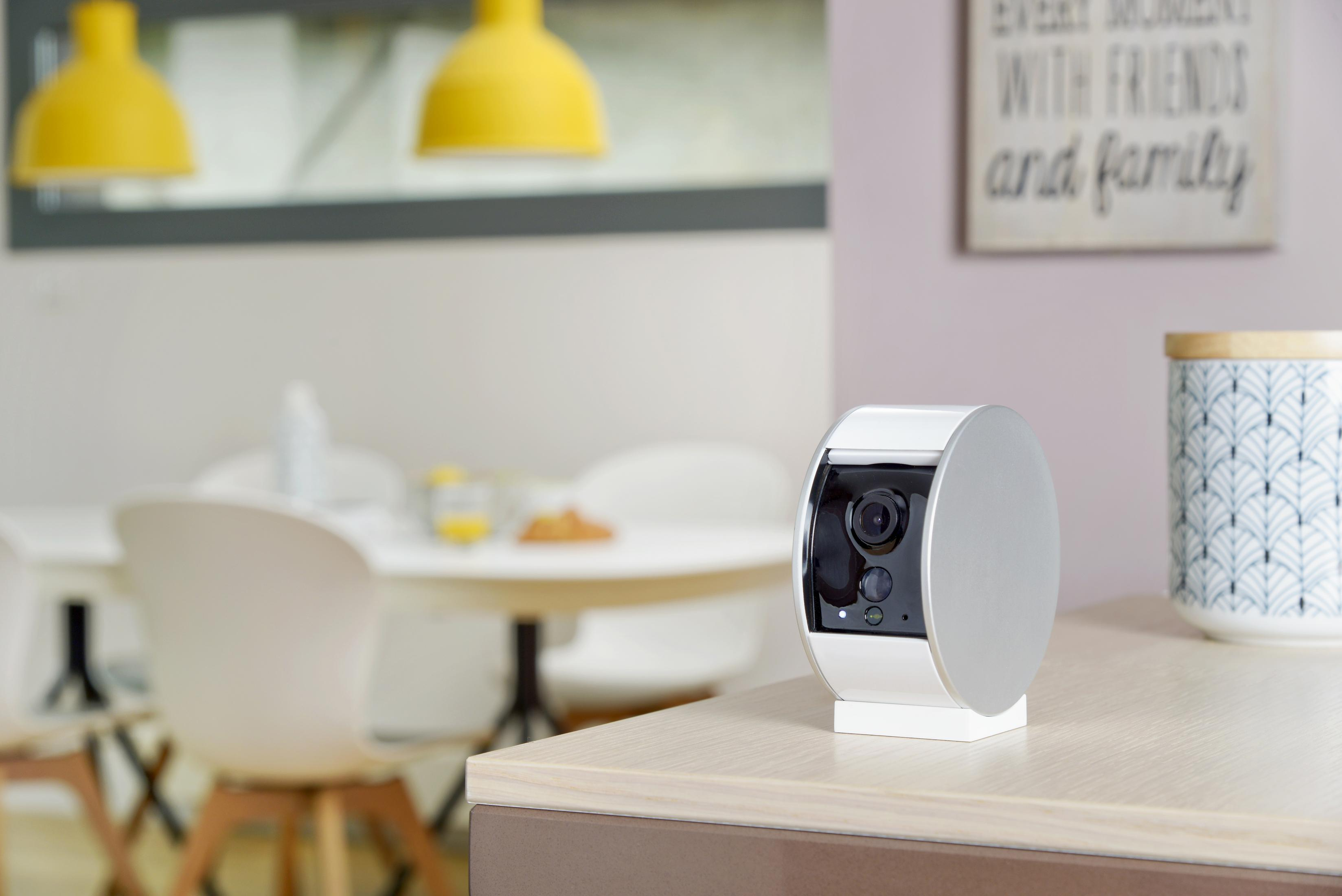 Die Arlo Überwachungskamera steht auf einer Holzoberfläche mit einem Essbereich im Hintergrund.