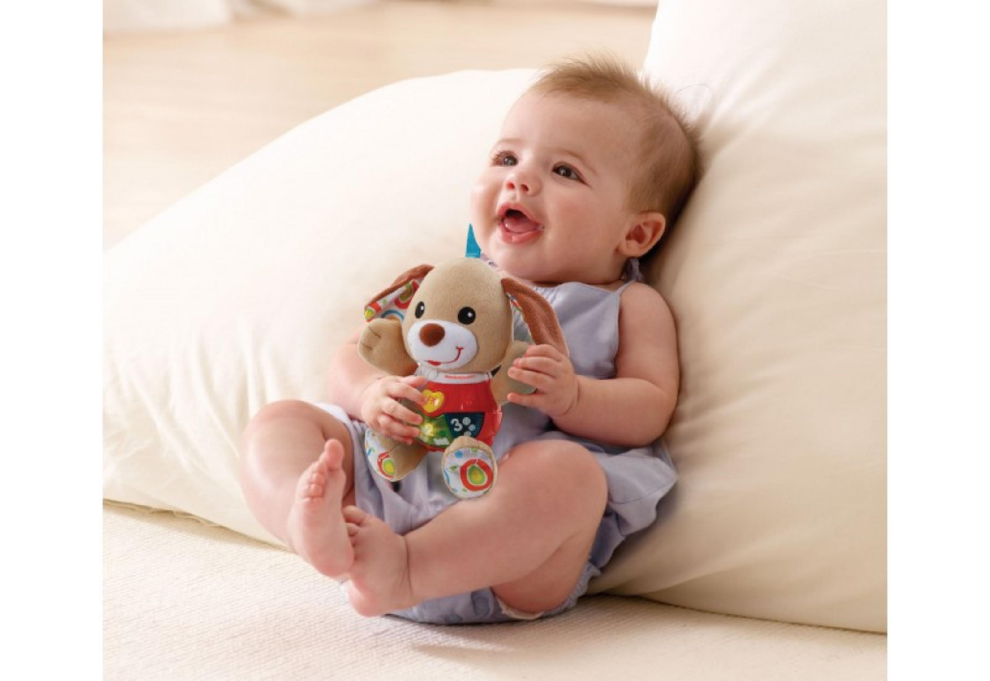 Ein Baby in Blau, das ein Plüschhundespielzeug mit bunten Akzenten hält, sitzt vor einem weißen Kissen, freudiger Ausdruck.