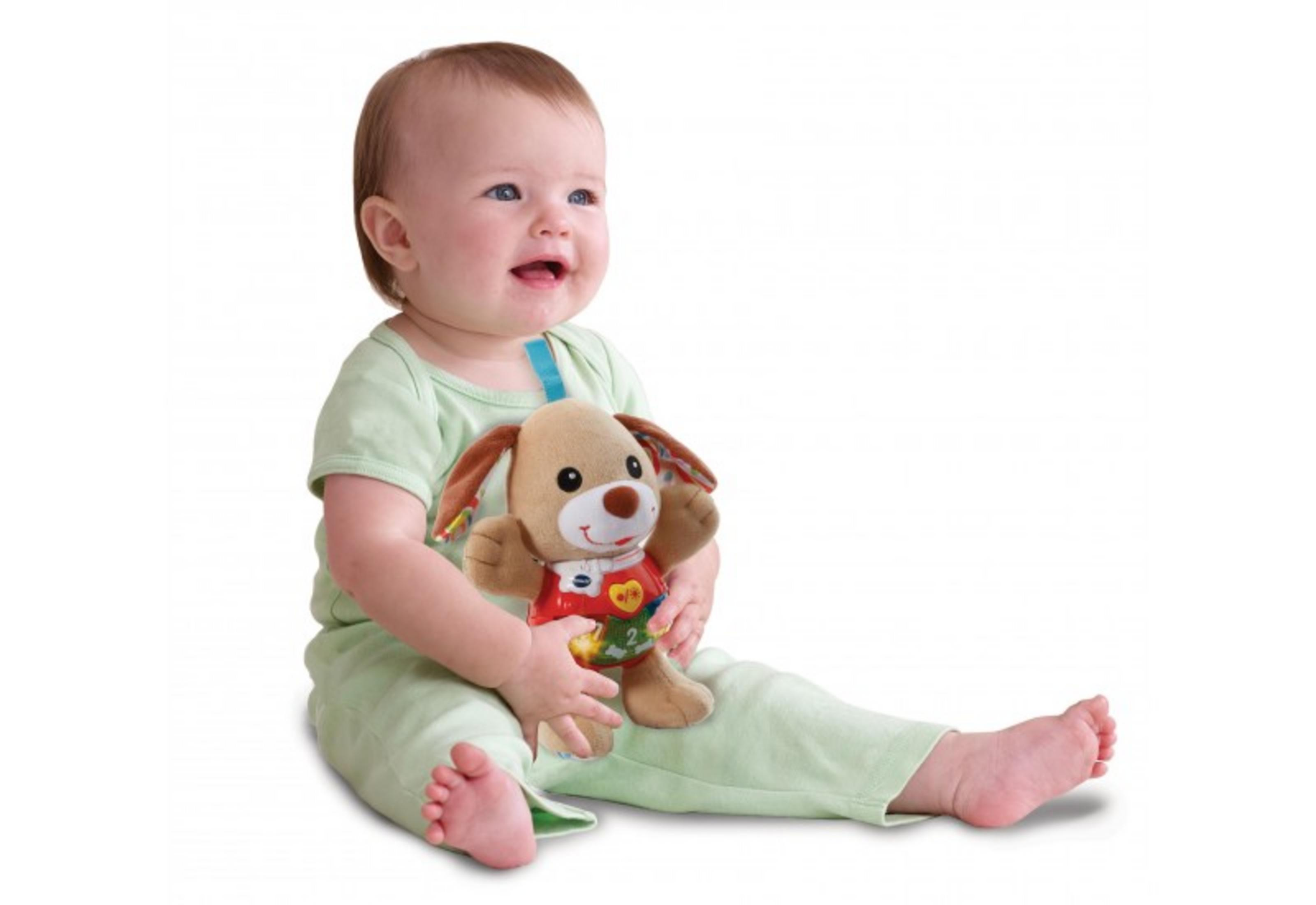 Ein Baby in Grün, das ein Plüschhundespielzeug mit bunten Akzenten hält, sitzt vor einem weißen Hintergrund, freudiger Ausdruck.