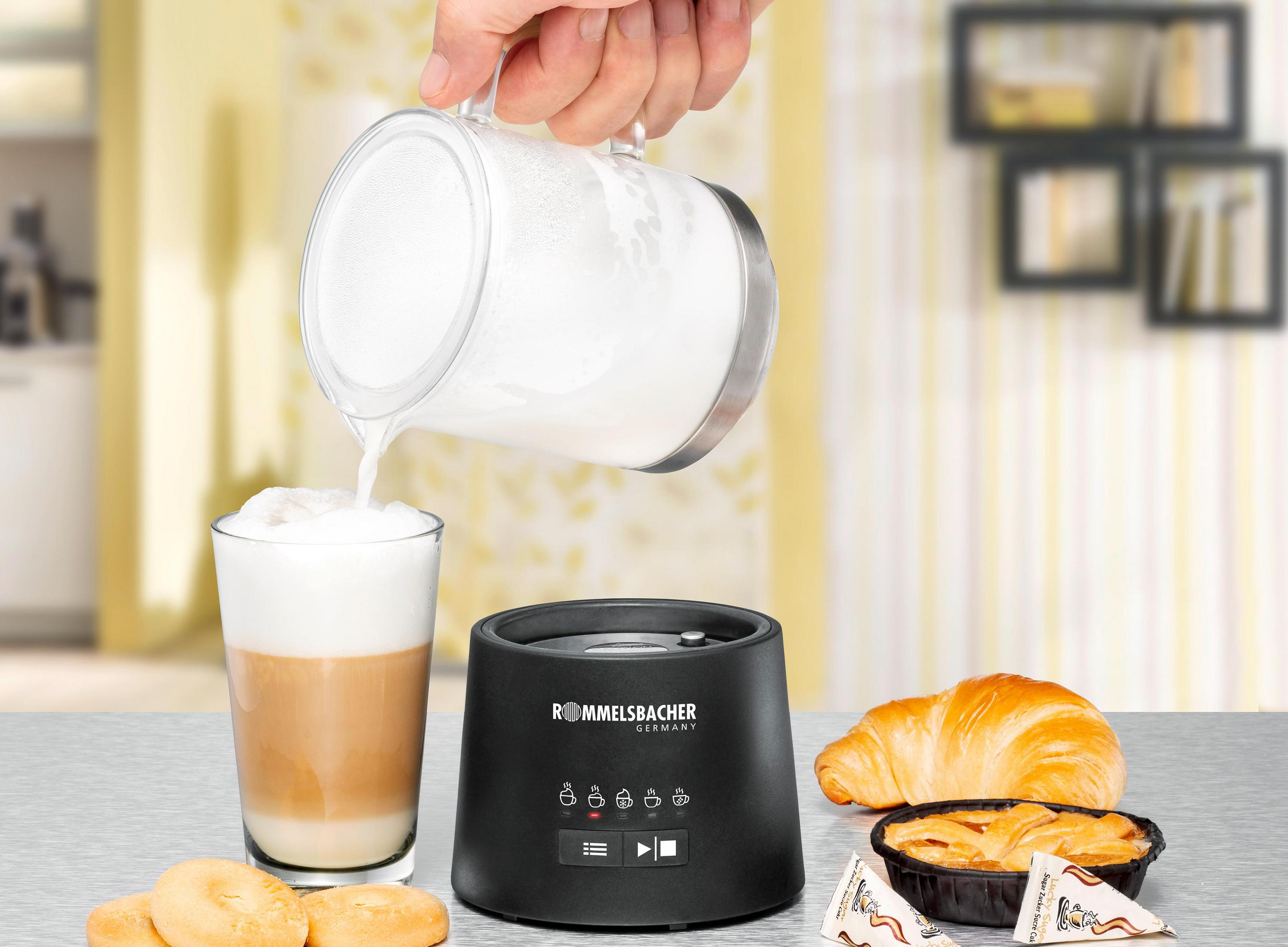 Eine Hand gießt Milch in einen Latte neben einem Milchaufschäumer und Gebäck.