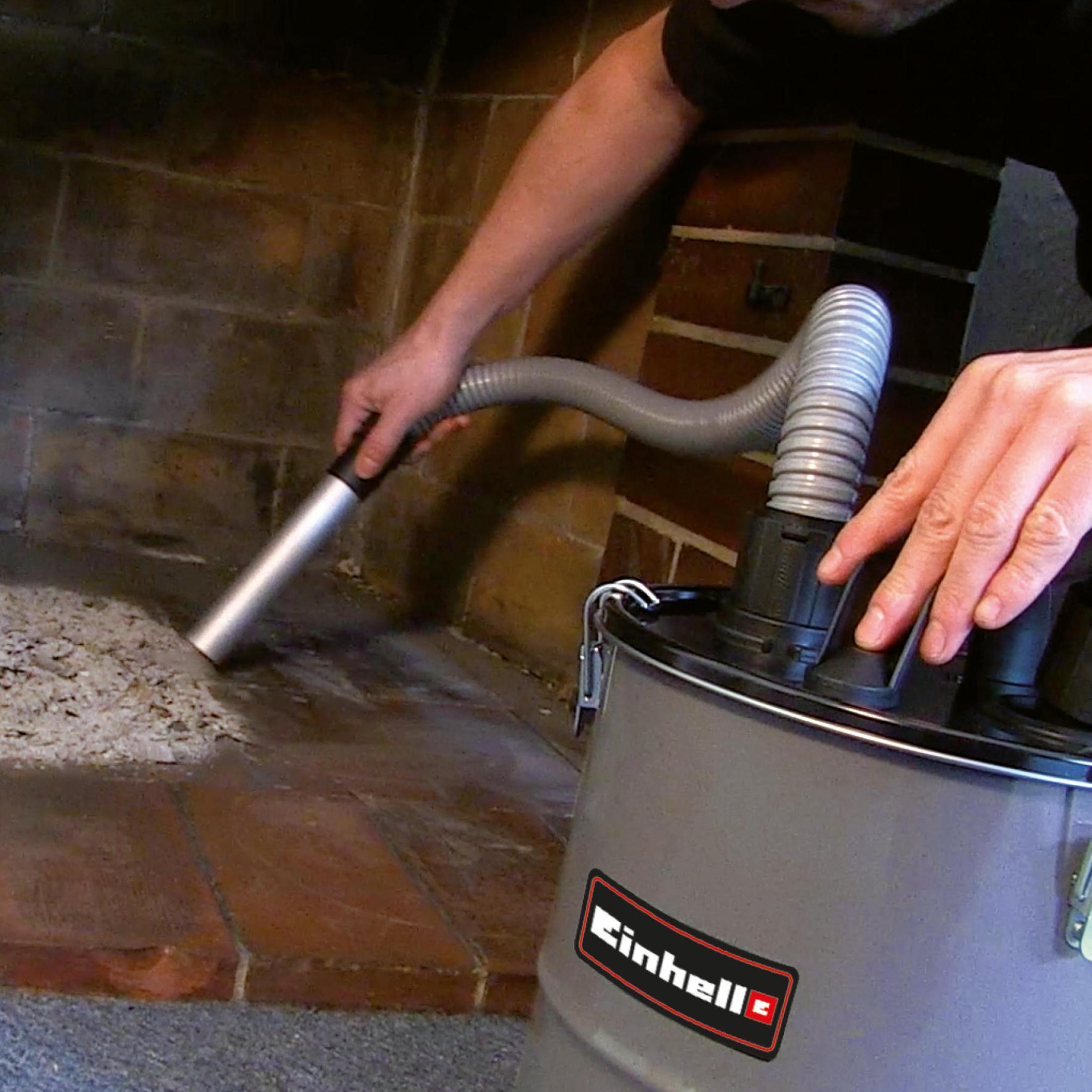 Eine Person benutzt einen grauen Einhell Aschesauger, um einen mit weißer Asche bedeckten Kamin aus Ziegeln zu reinigen. Fokus auf Einhell-Produkt.