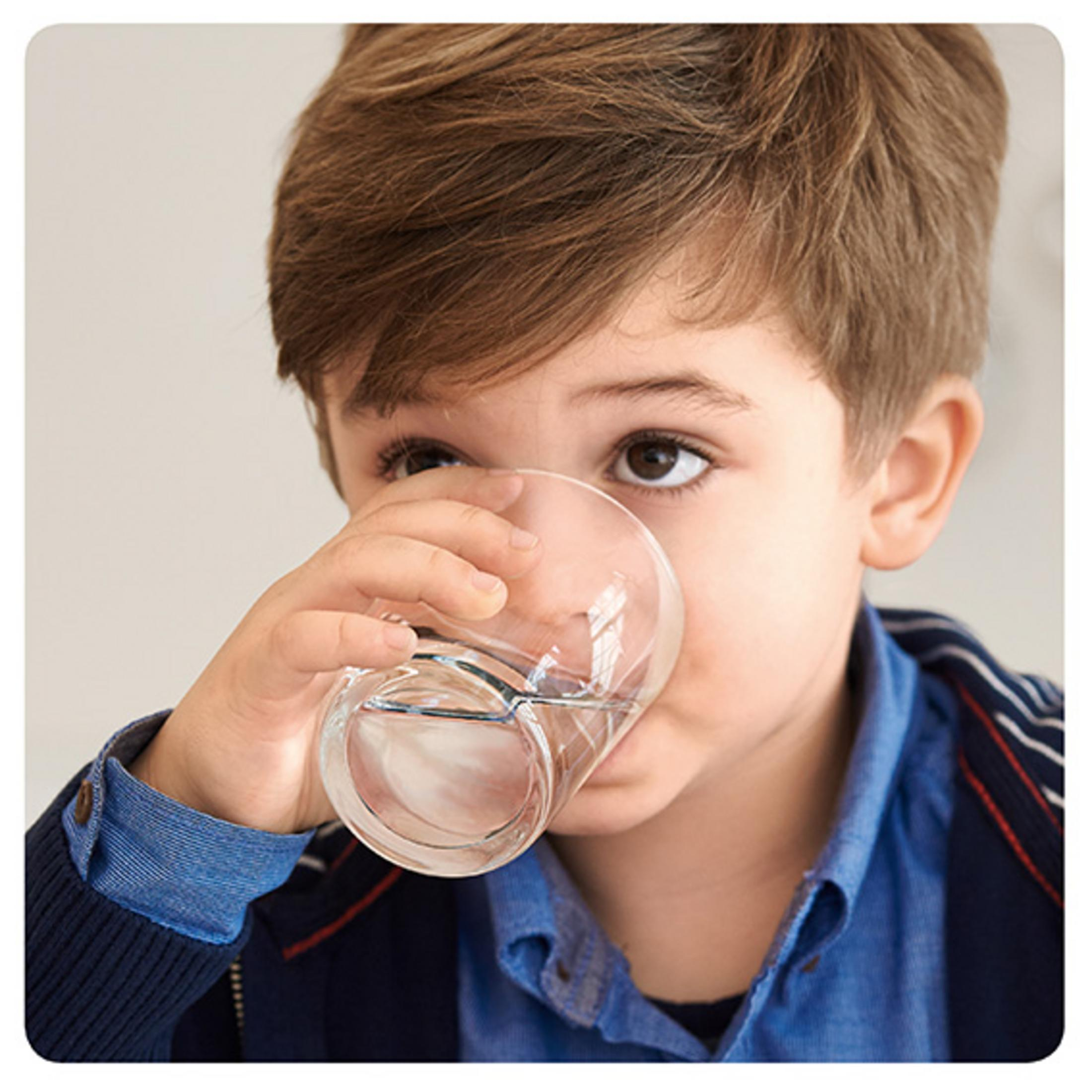 Ein junges Kind trinkt Wasser aus einem klaren Glas und trägt ein blaues Button-Down-Hemd.