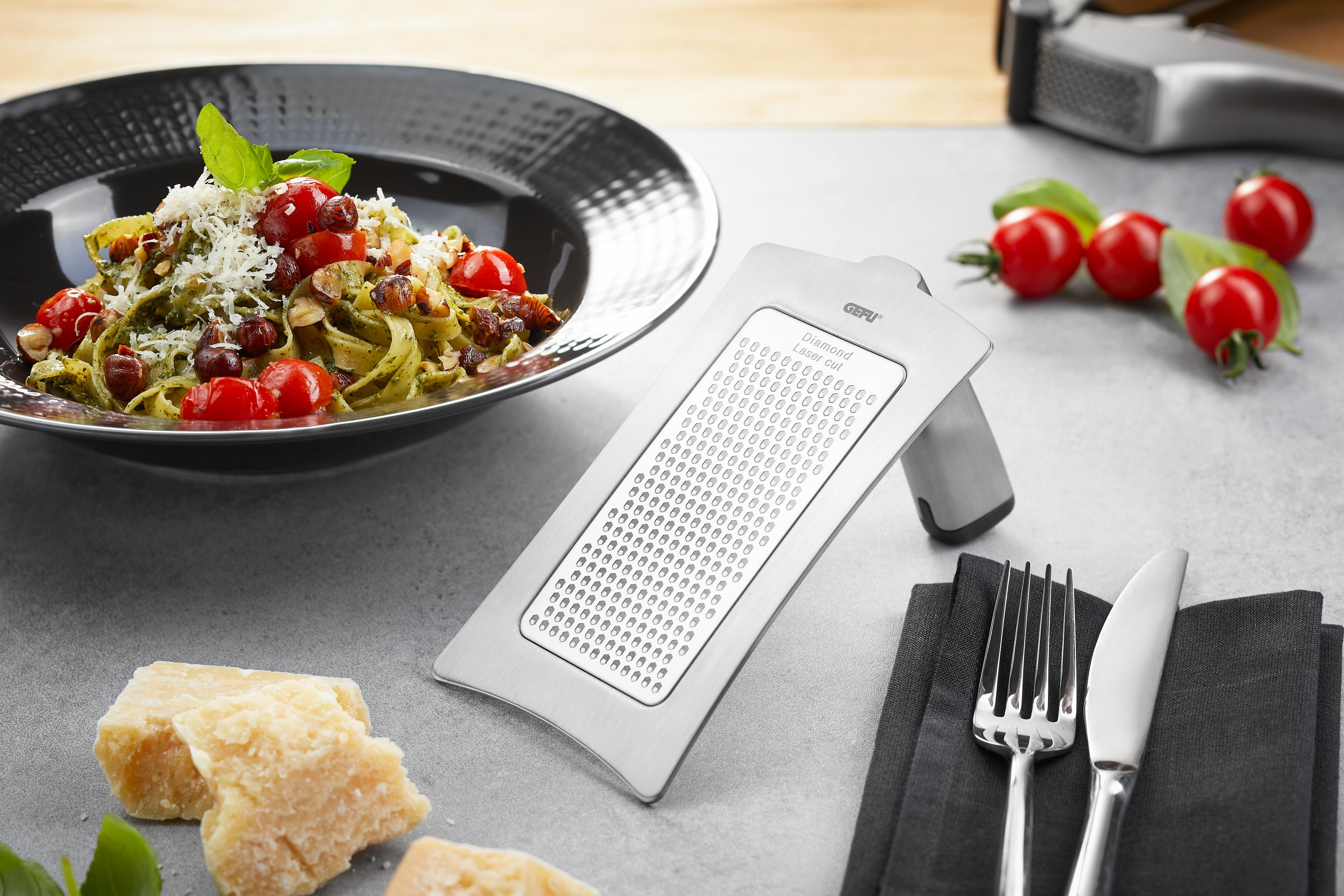 GEFU-Reibe mit Nudelgericht, Tomaten, Parmesan, Besteck und einer schwarzen Serviette, die die Zubereitung von Gourmetgerichten zeigt.