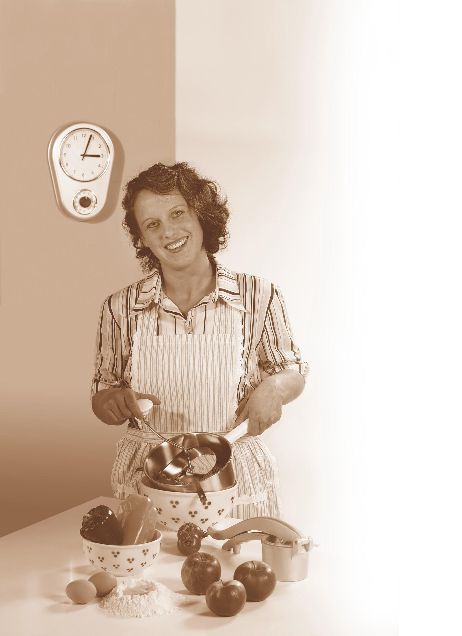 Eine Frau mit lockigem Haar lächelt, während sie eine Gemüsemühle benutzt, umgeben von Küchenutensilien und Zutaten.