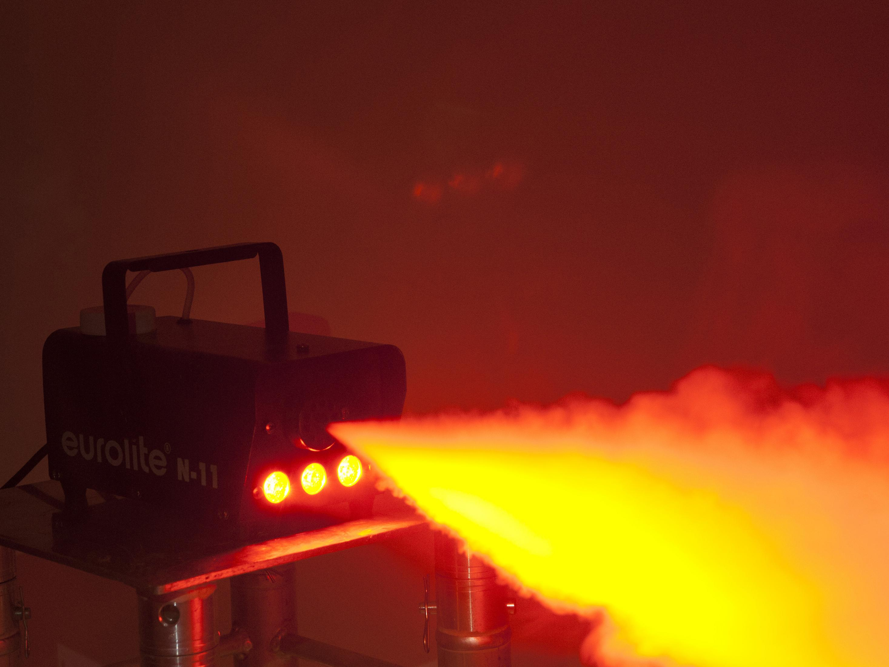 Eurolite N-11 Nebelmaschine, die gelben Rauch in einem rot beleuchteten Raum erzeugt. Die Maschine steht auf einem Metallständer.