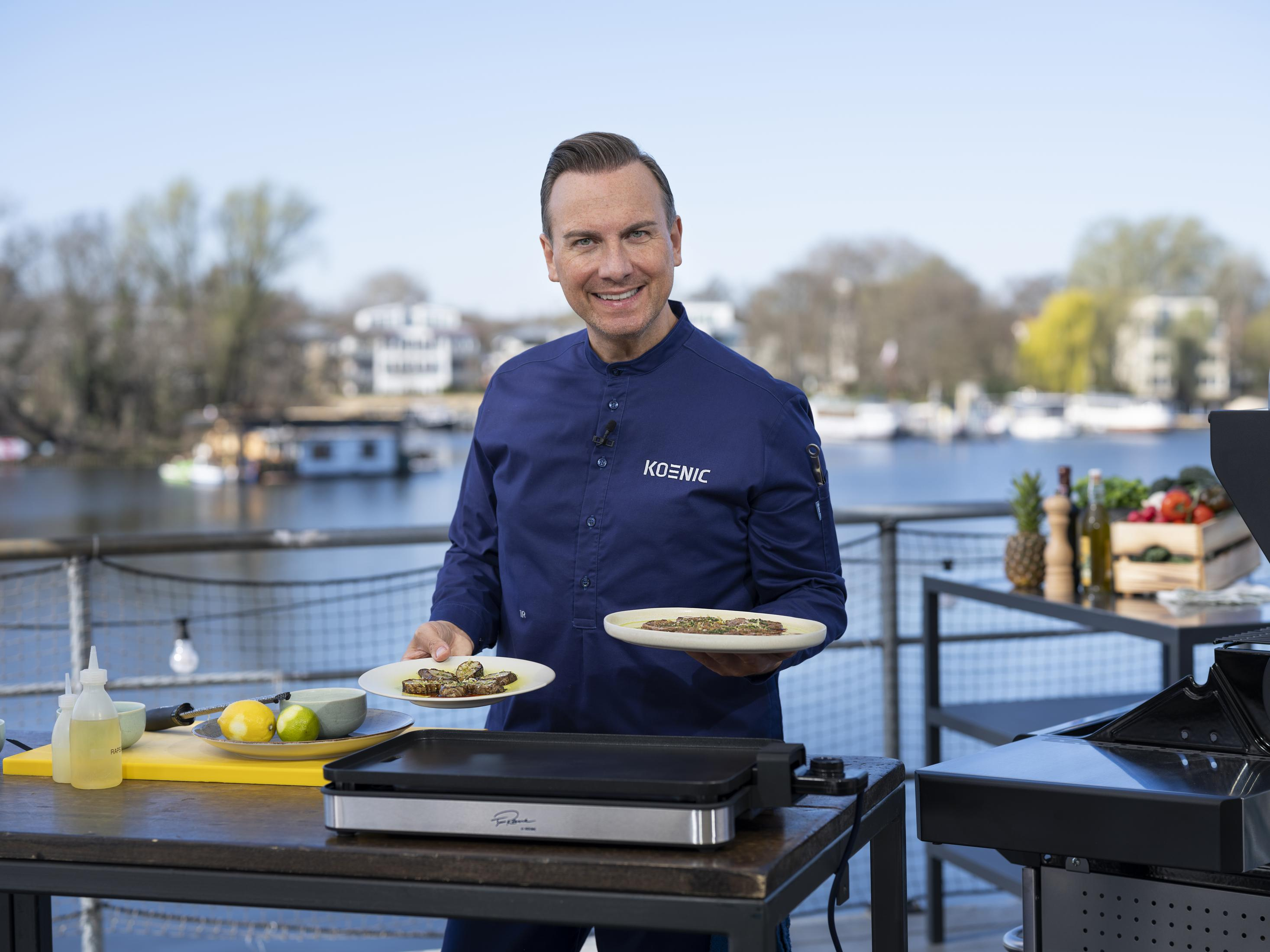 Ein Mann hält Teller mit Essen vor einem Grill auf einer Außenterrasse.