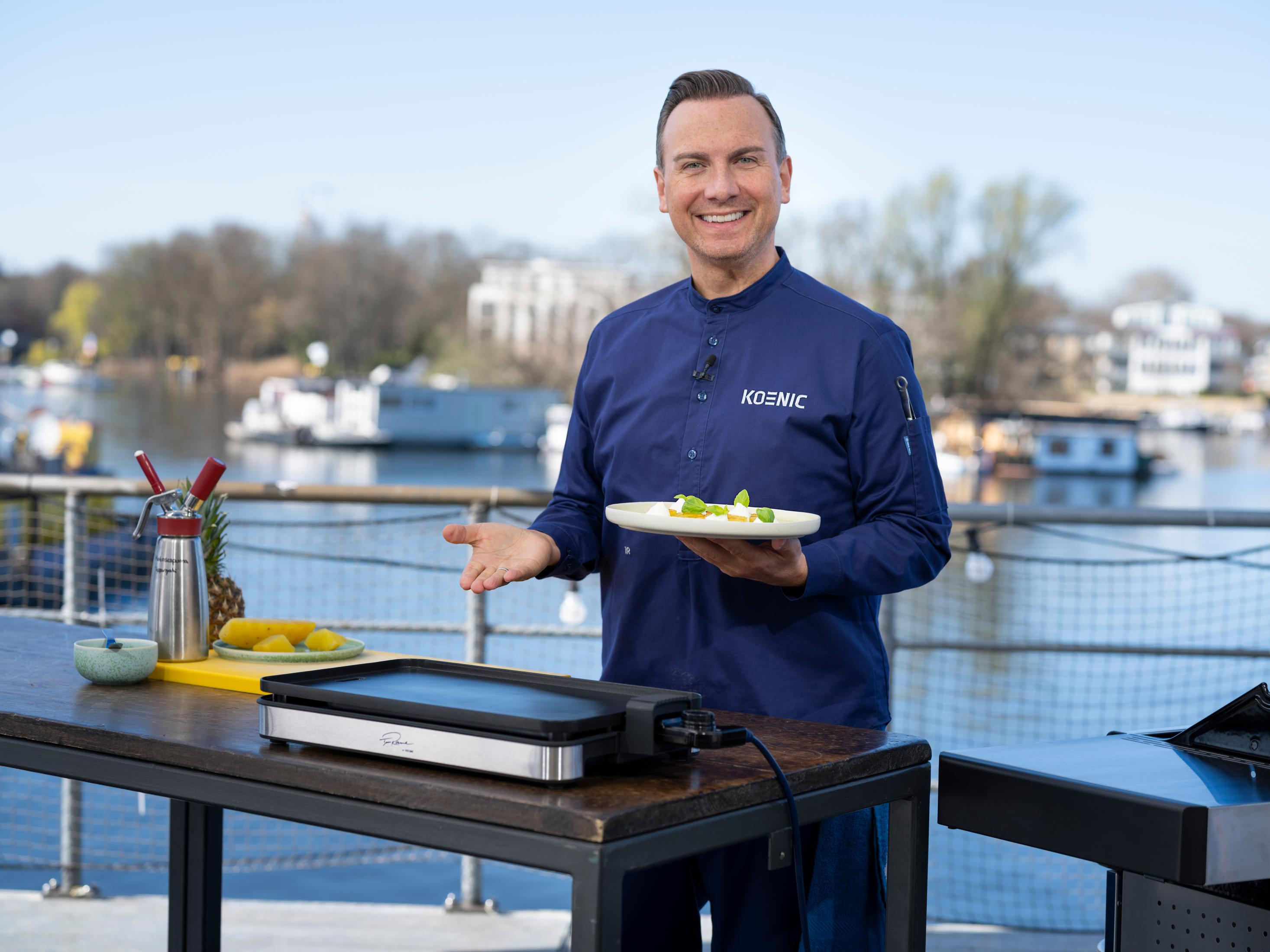 Ein Mann hält einen Teller mit Essen vor einem Grill auf einer Außenterrasse.