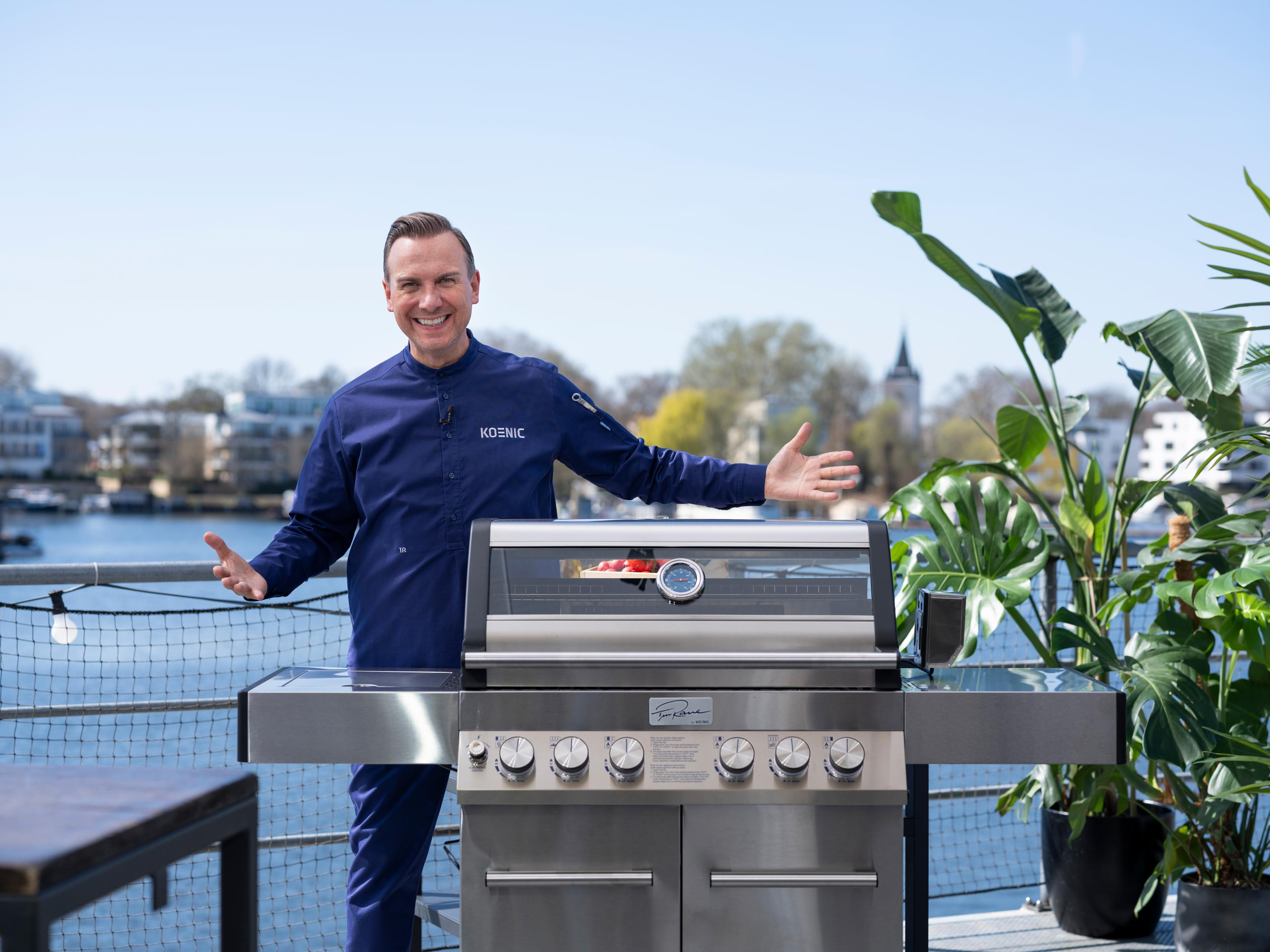 Ein lächelnder Mann steht mit ausgestreckten Armen hinter einem Edelstahlgrill vor einem sonnigen Hintergrund im Freien.