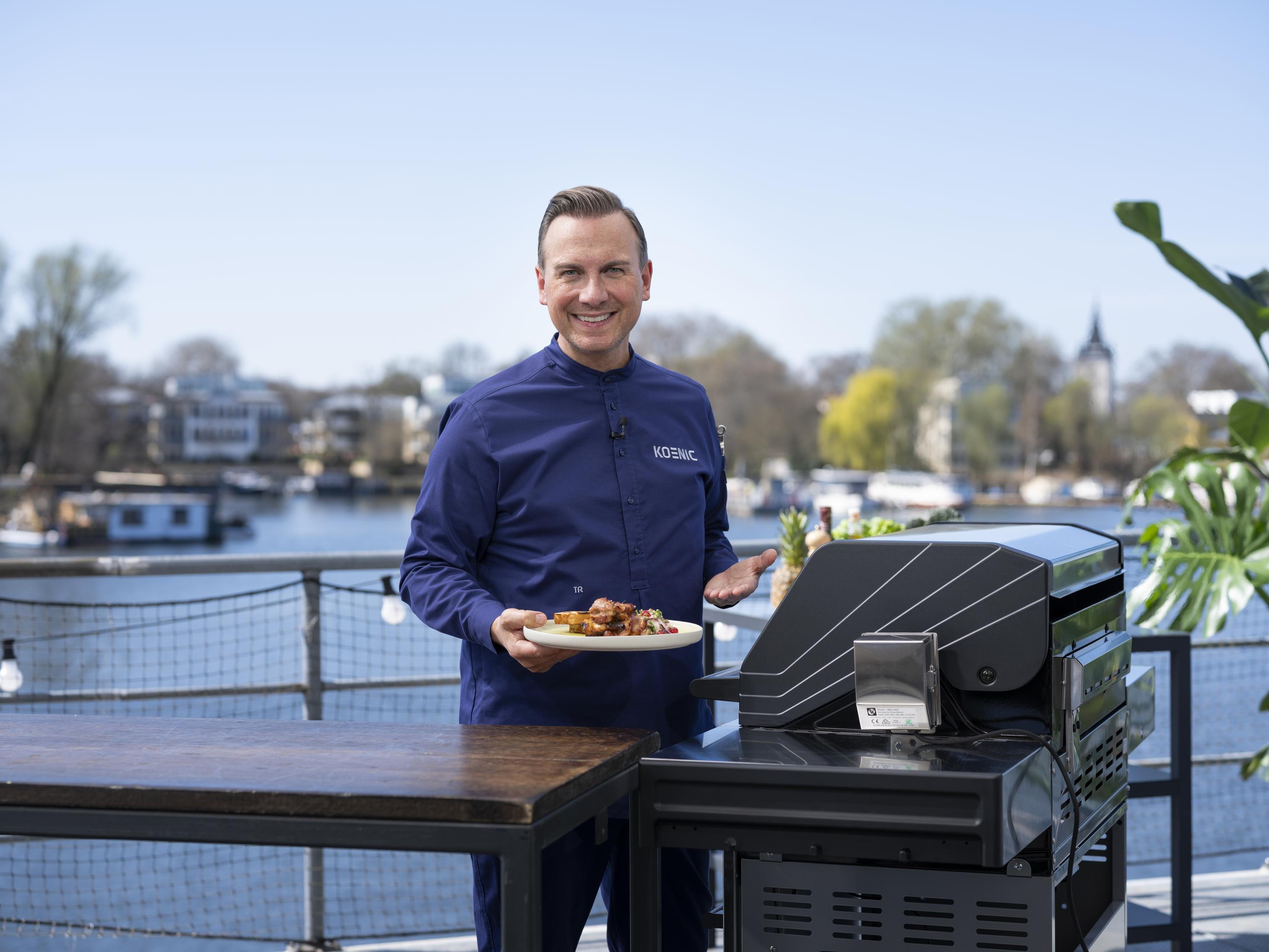 Lächelnder Mann, der einen Teller mit Essen neben einem Grill an einem sonnigen Tag im Freien hält.