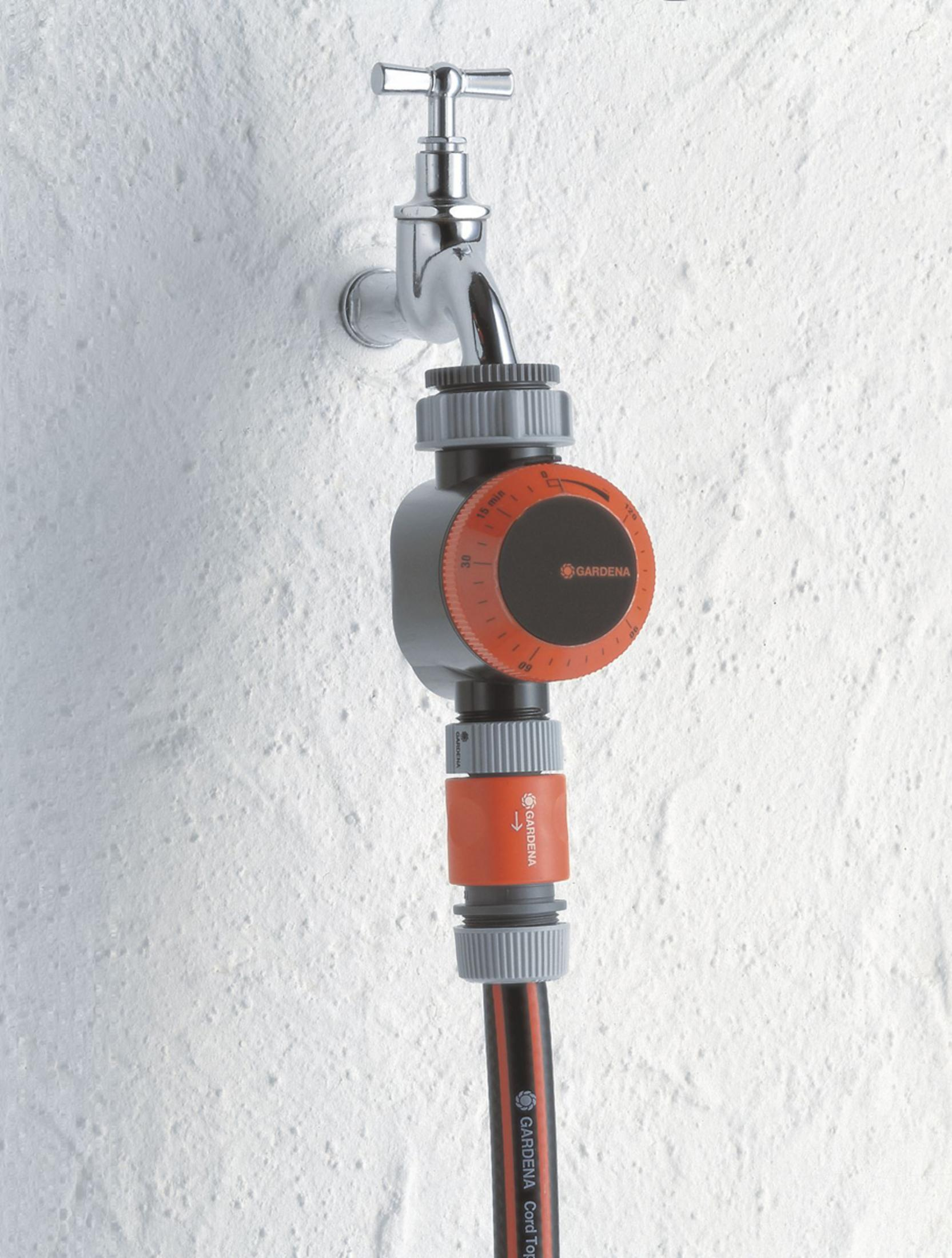 Ein grauer Gardena-Wasserzeitschalter ist an einem Wasserhahn an einer weißen Wand befestigt, wobei darunter ein Gardena-Schlauch angeschlossen ist.