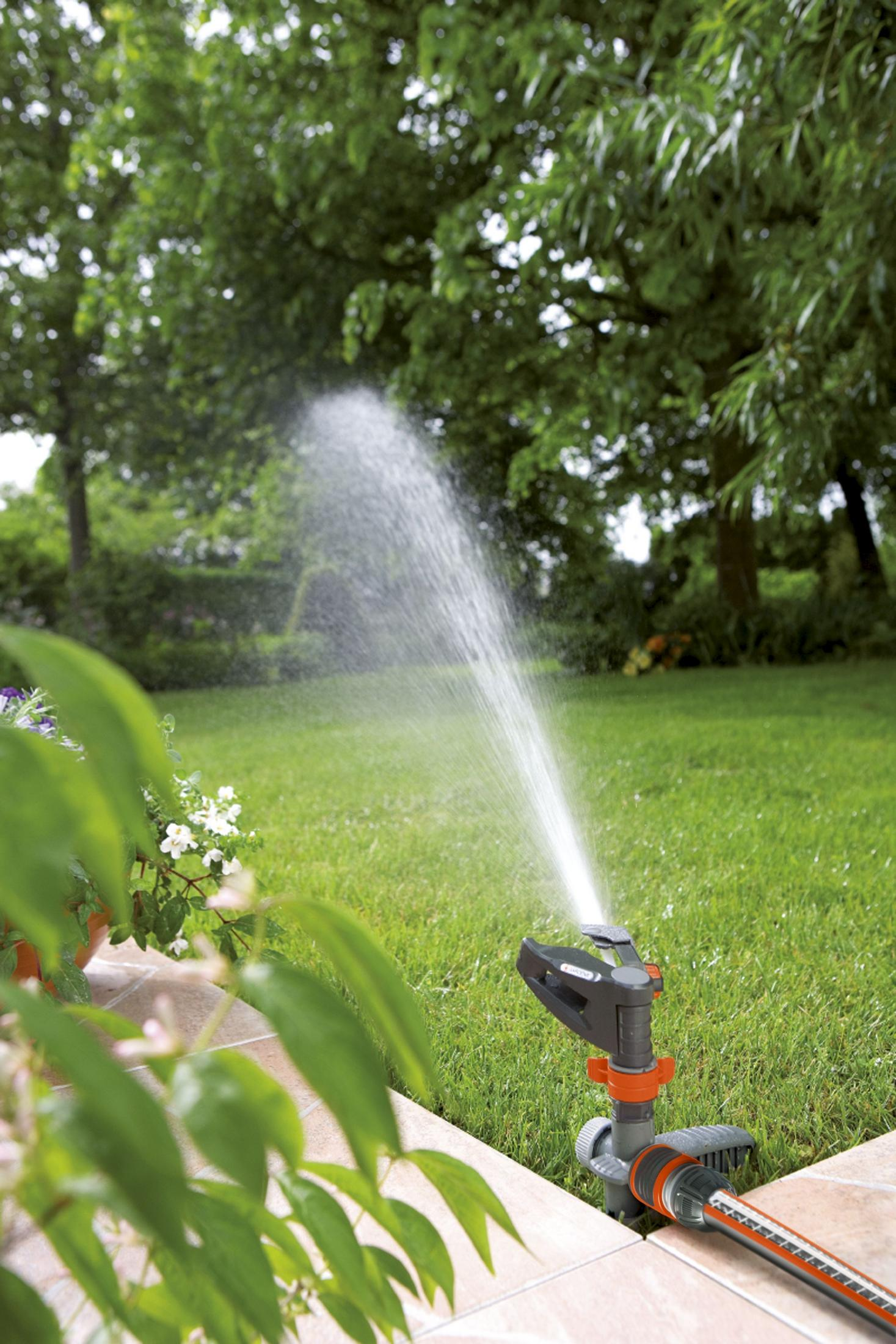 Ein Gartensprenger sprüht Wasser auf einen grünen Rasen, mit Bäumen und Blumen im Hintergrund.