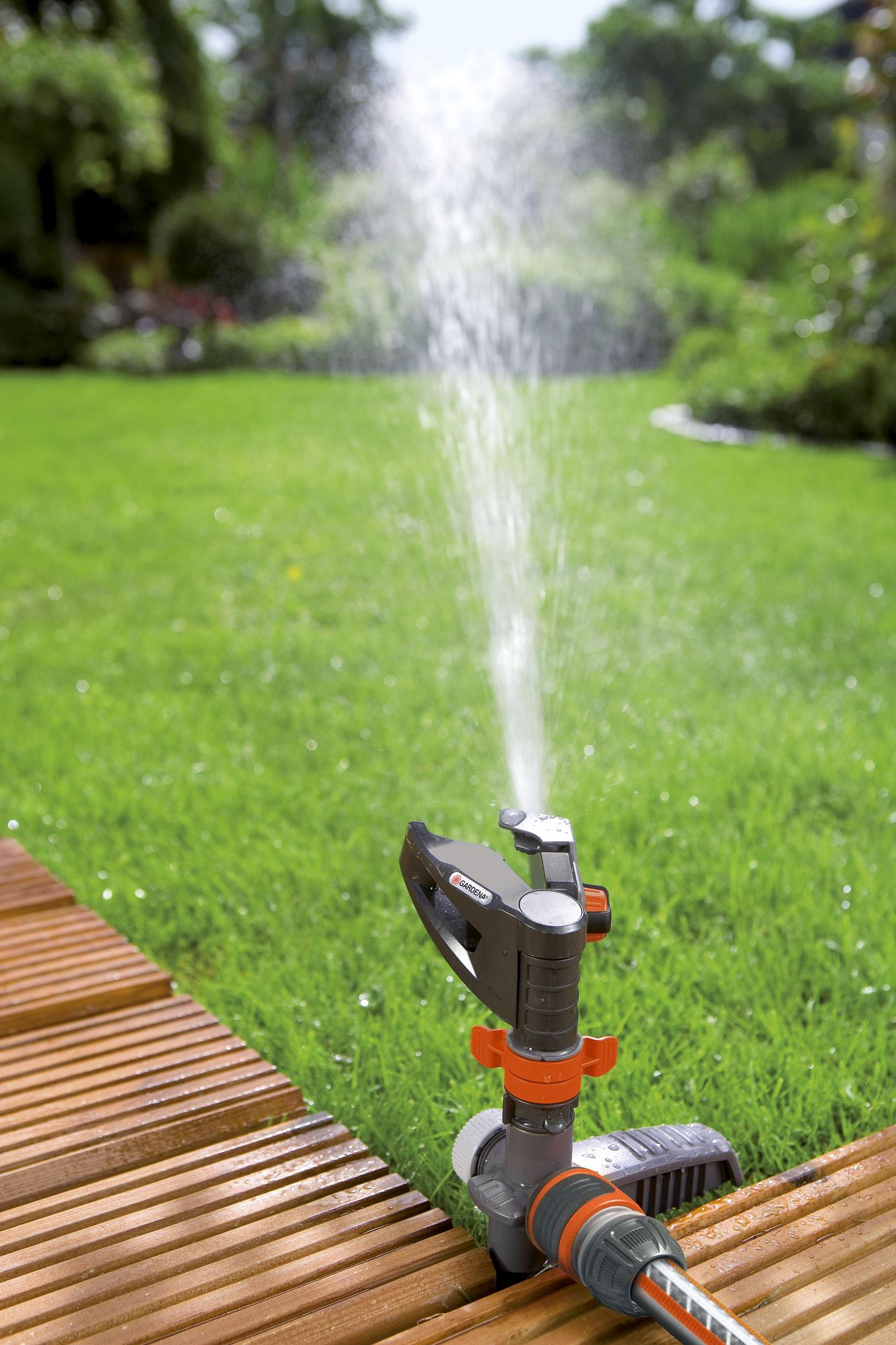 Ein Gartensprenger sprüht Wasser auf einen grünen Rasen, positioniert auf einem Holzdeck.