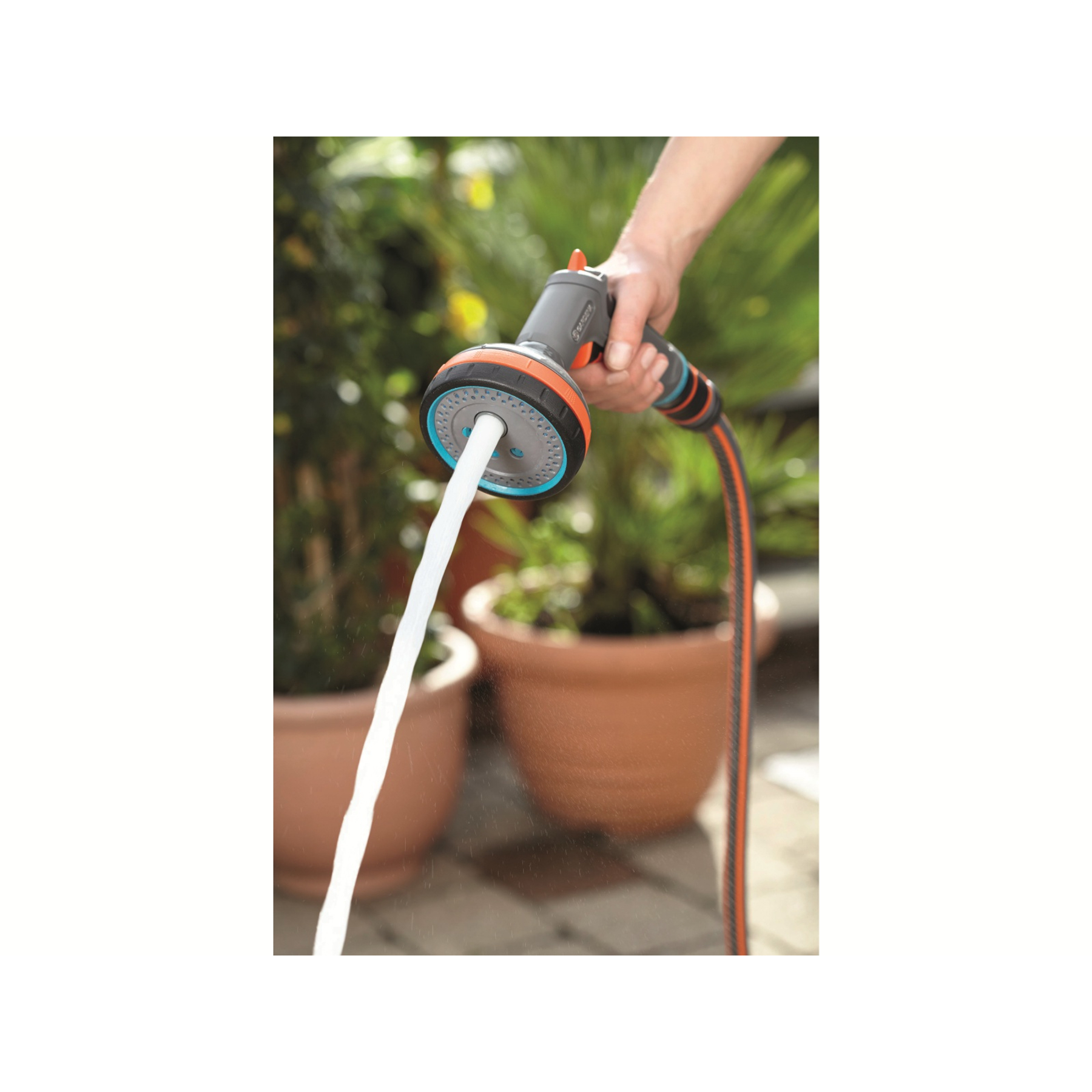 Una mano tiene un ugello di spruzzo Gardena grigio e arancione che spruzza acqua, con vasi di fiori sullo sfondo.