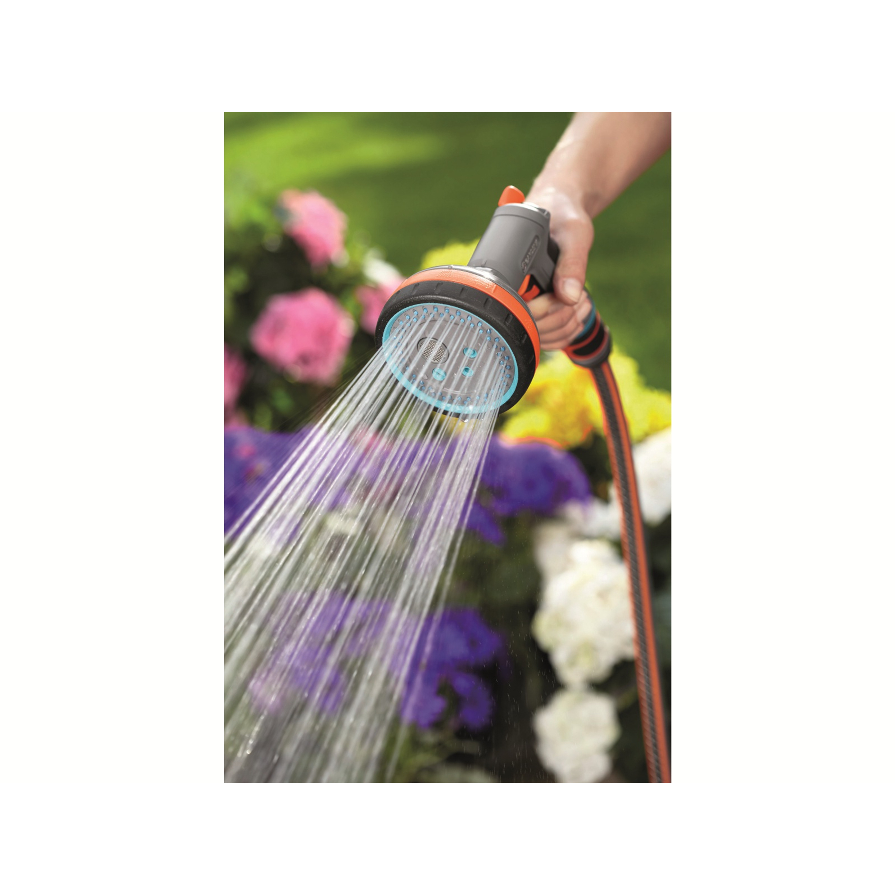 Una mano tiene un ugello di spruzzo Gardena grigio e arancione che spruzza acqua, con fiori sullo sfondo.