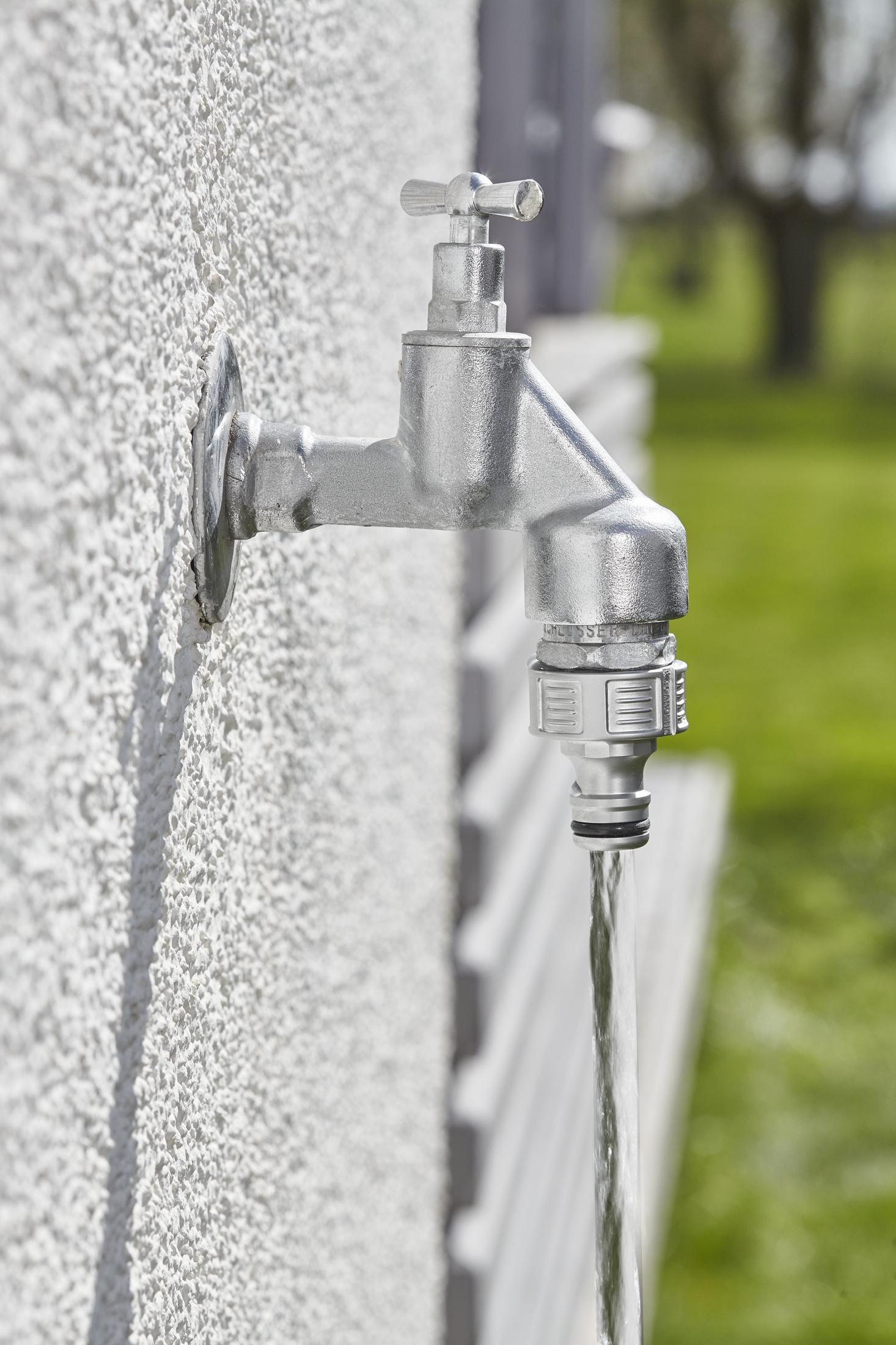 Rubinetto argentato fissato a un muro bianco, con acqua che scorre dal connettore del tubo Gardena.