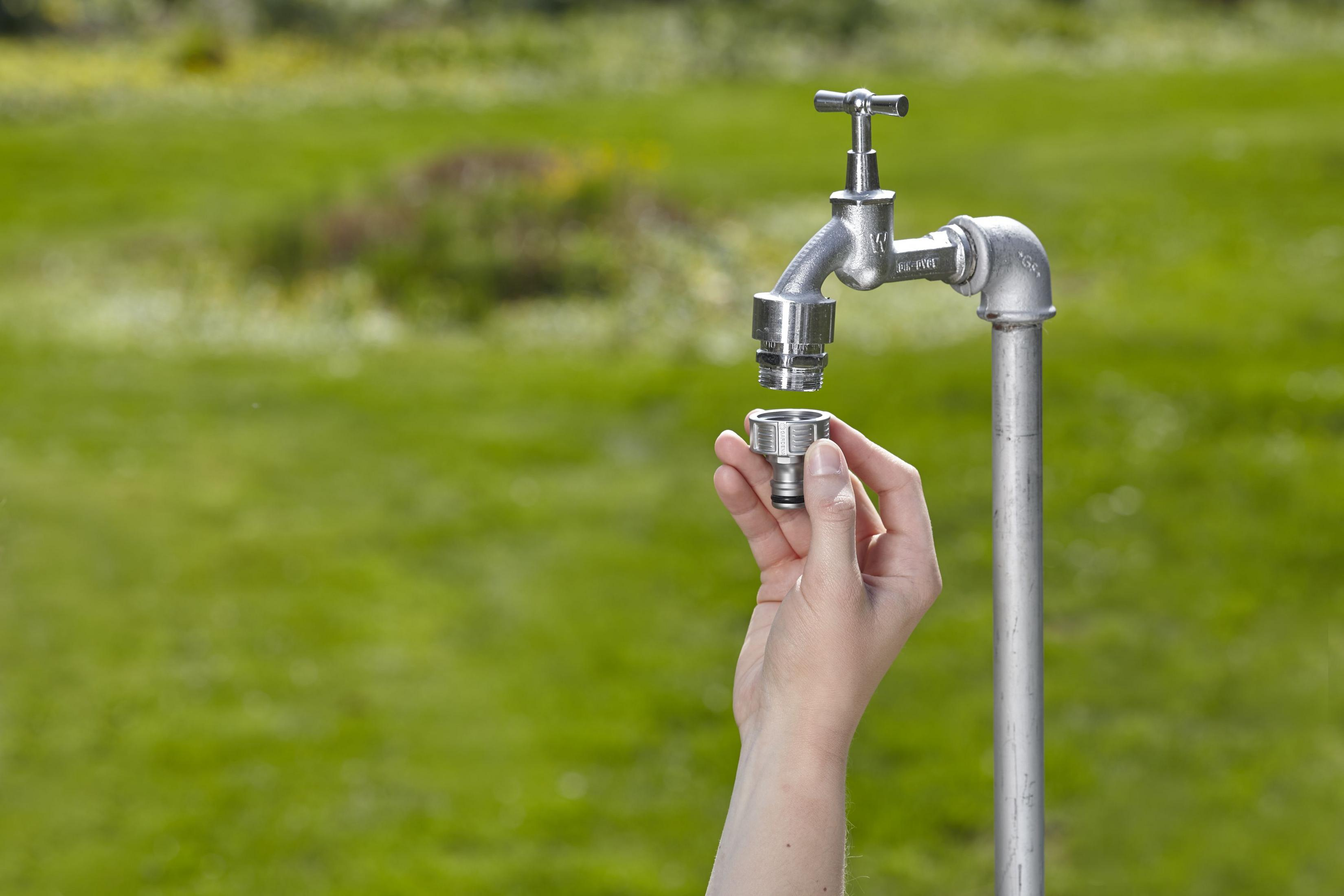 Una mano che tiene un connettore per tubo Gardena argentato vicino a un rubinetto, con un campo verde sullo sfondo.