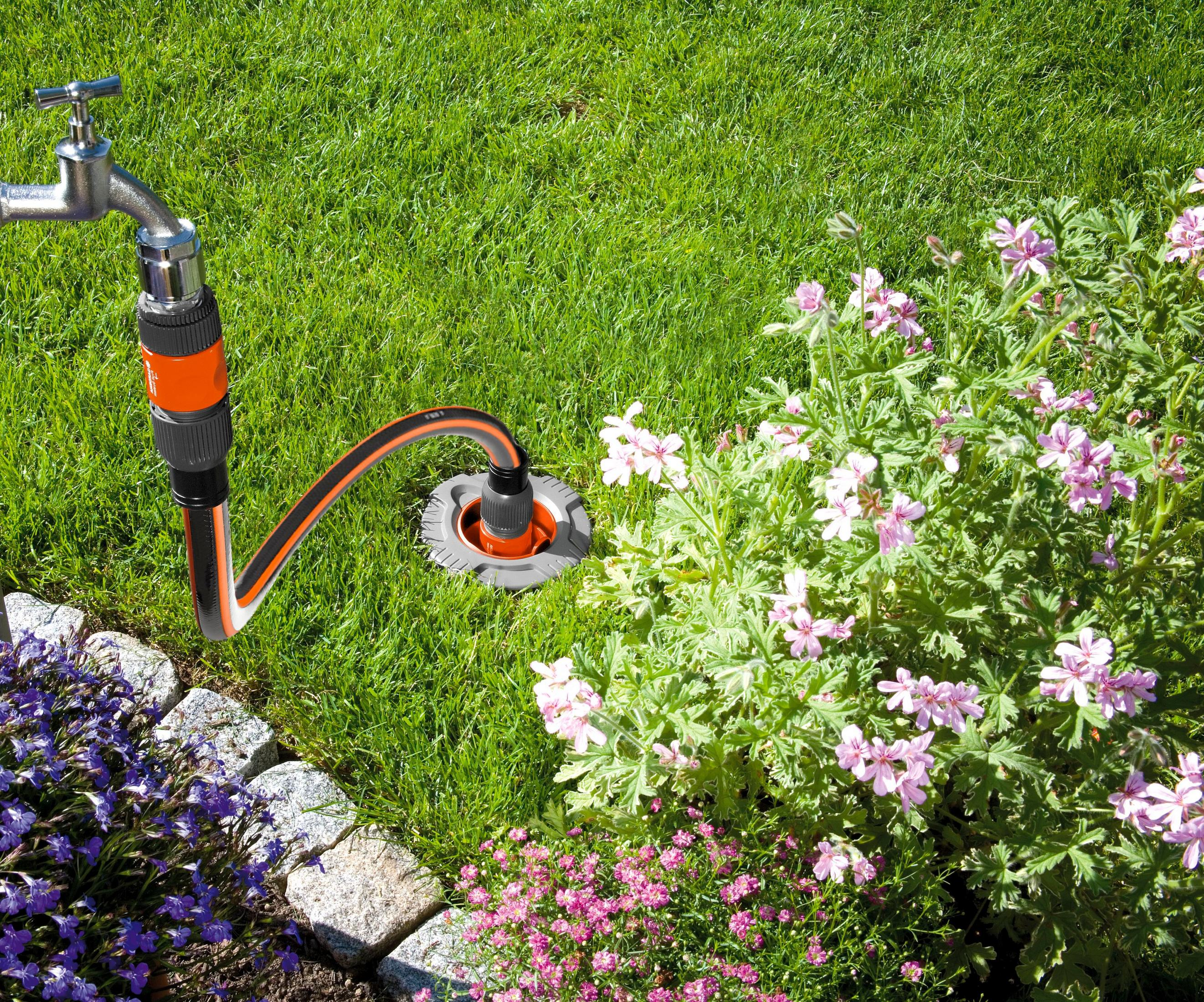 Ein Schlauch, der an einem Wasserhahn angeschlossen ist, bewässert Blumen auf einem grünen Rasen mit Steinrand.