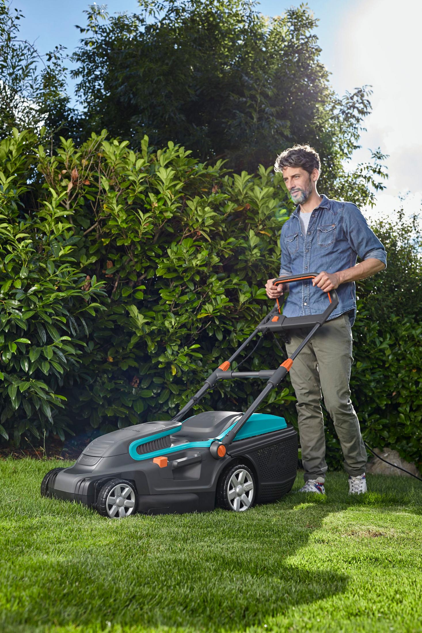 Ein Mann in einem Jeanshemd mäht den Rasen mit einem grauen und teal Rasenmäher.