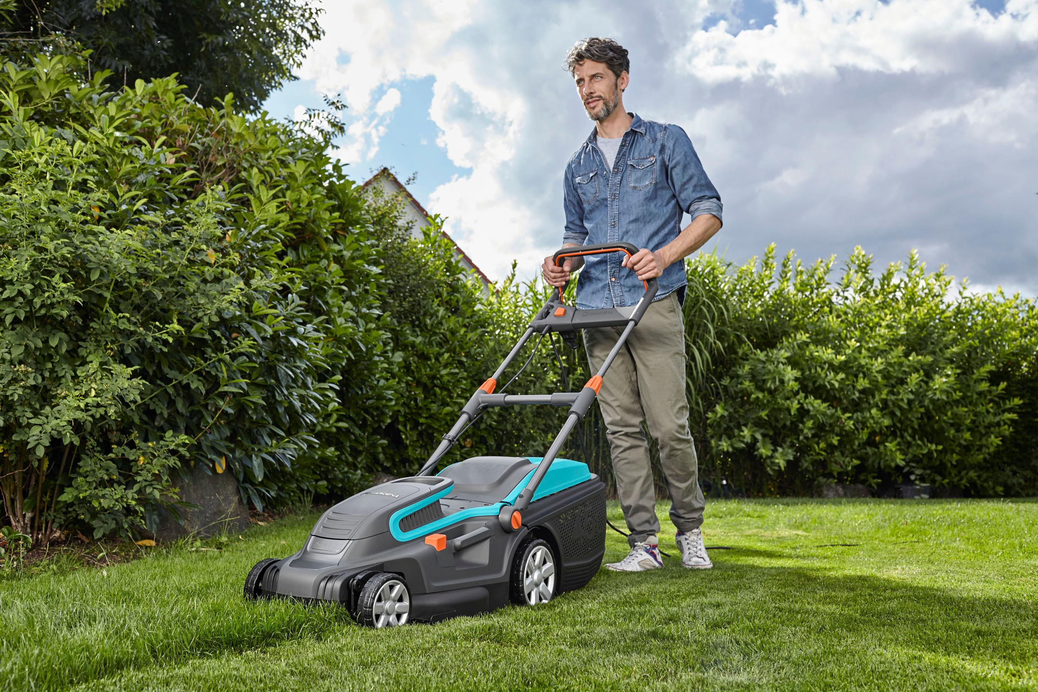 Ein Mann in einem Jeanshemd schiebt einen grauen und teal Rasenmäher auf einem grünen Rasen.