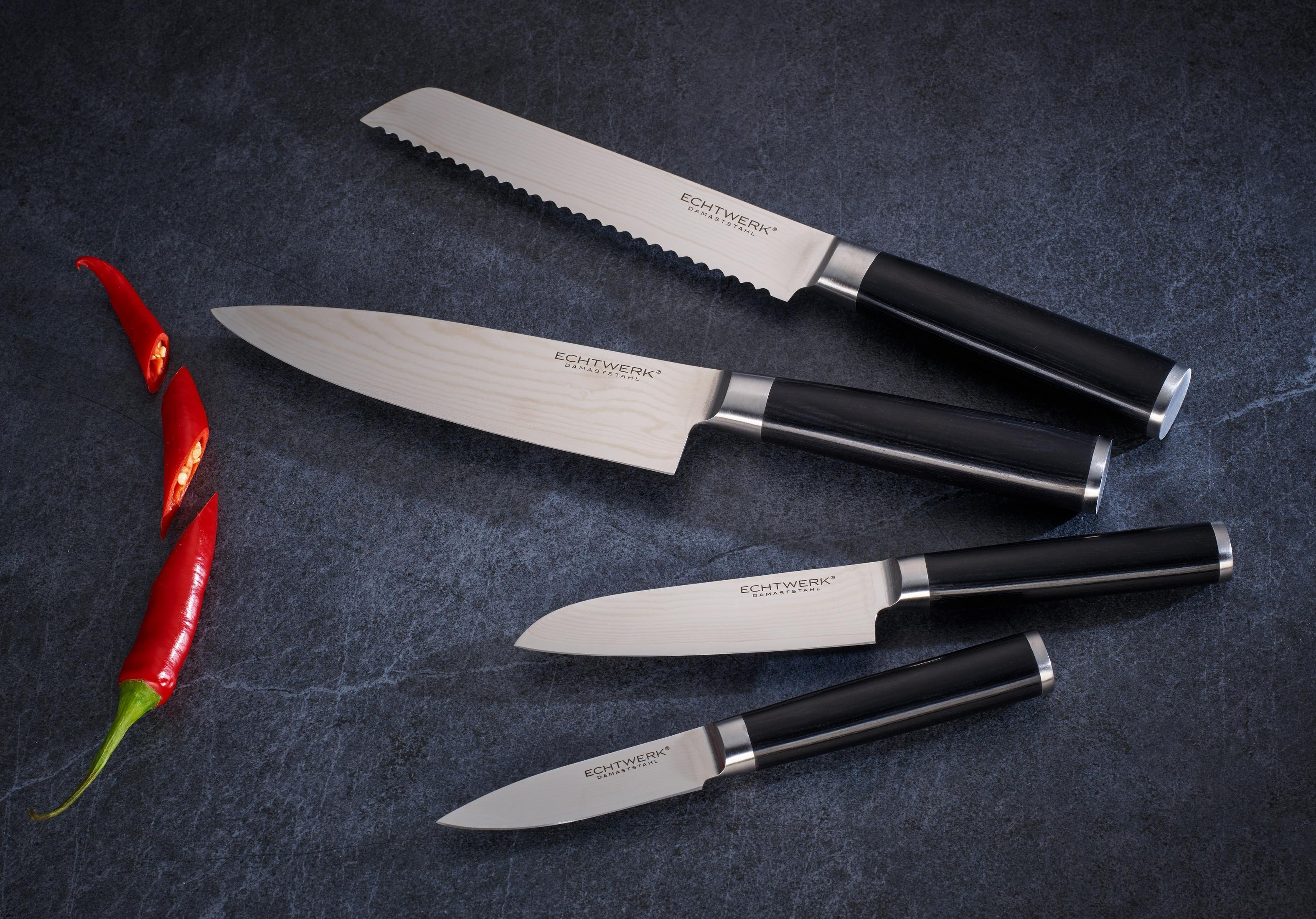 Vier Küchenmesser mit schwarzen Griffen und silbernen Klingen auf dunkler Oberfläche mit Chilischoten.