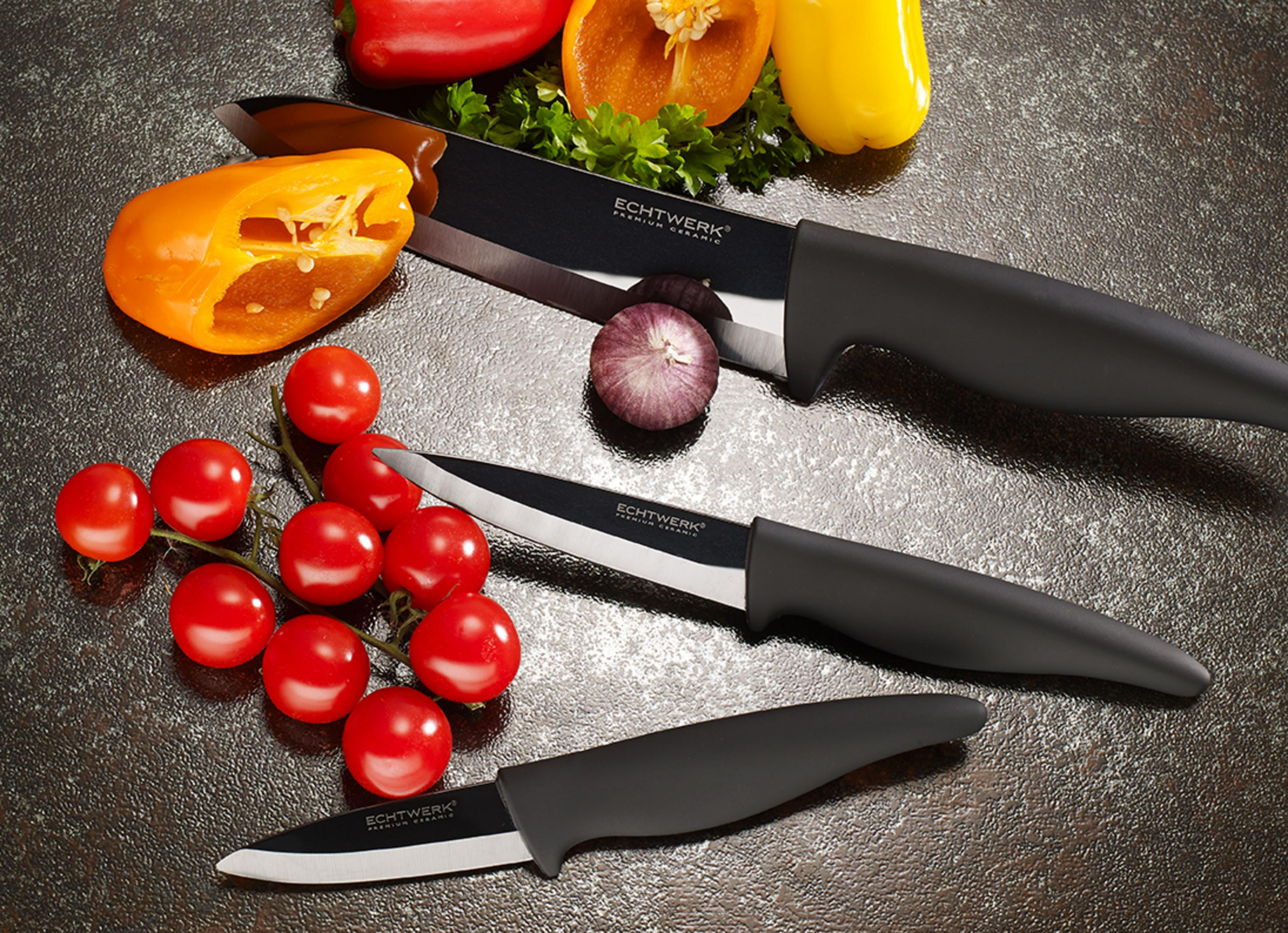 Drei schwarze Keramikmesser mit silbernen Klingen liegen auf einer dunklen Oberfläche mit Gemüse.