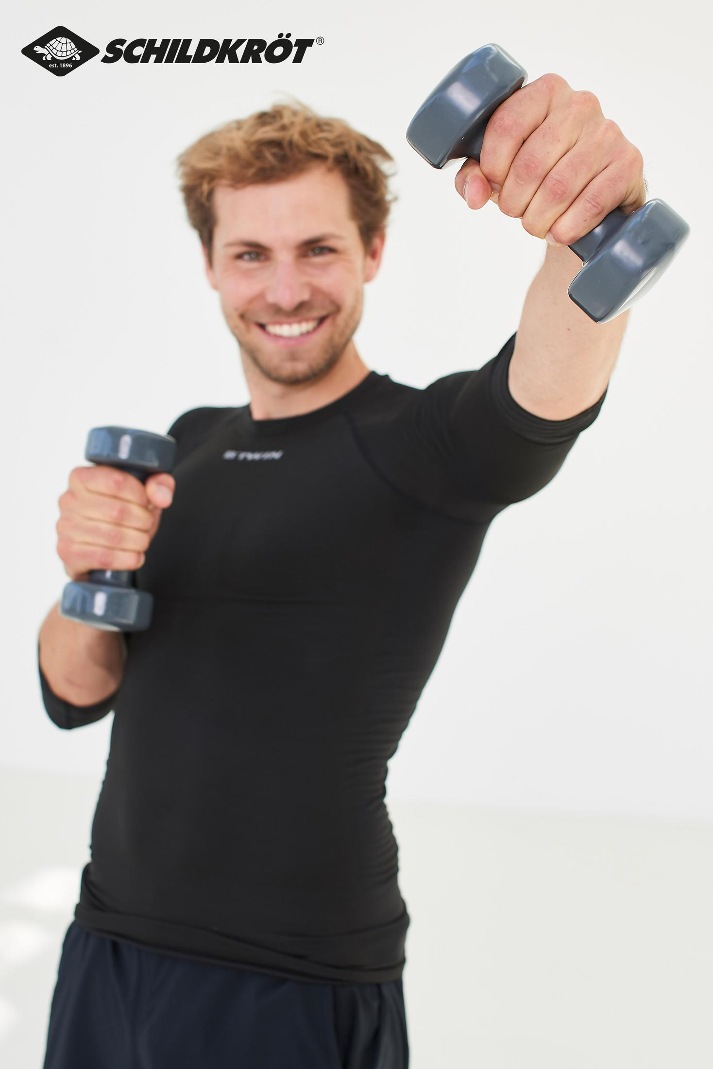 Un uomo sorridente in abbigliamento sportivo nero tiene in mano due manubri su uno sfondo bianco.
