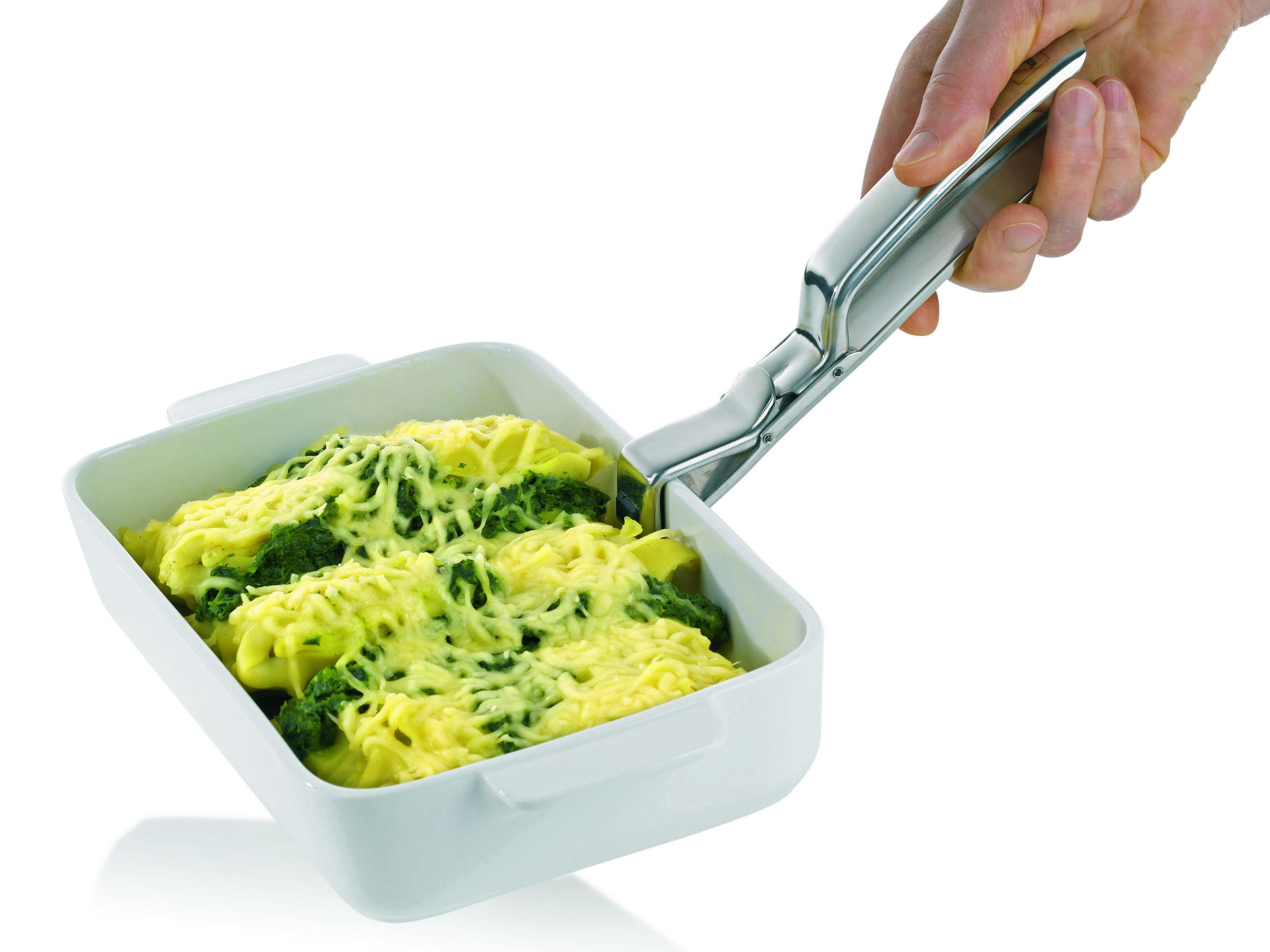 Ein silberner Pfannengreifer hält eine weiße Auflaufform, die mit käsiger Pasta und grünem Gemüse gefüllt ist.