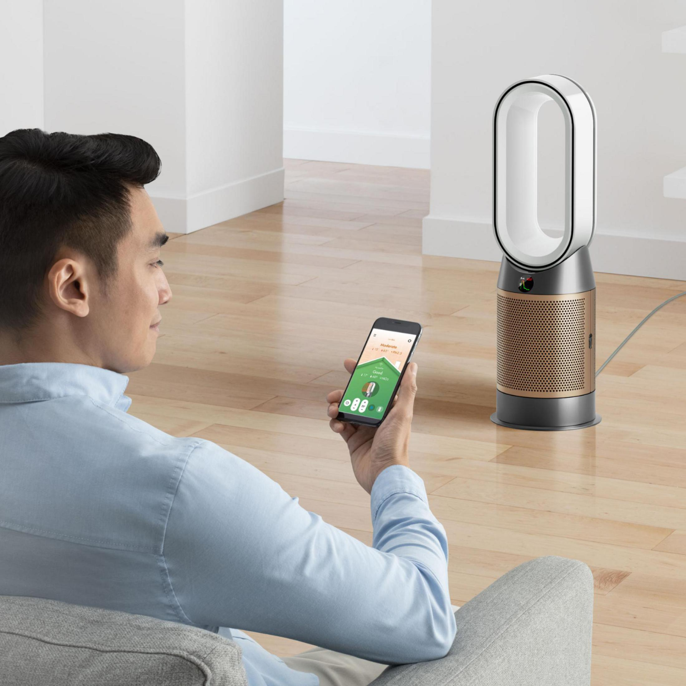 Un uomo usa il suo telefono vicino a un purificatore d'aria Dyson, seduto su un divano grigio in una stanza luminosa.