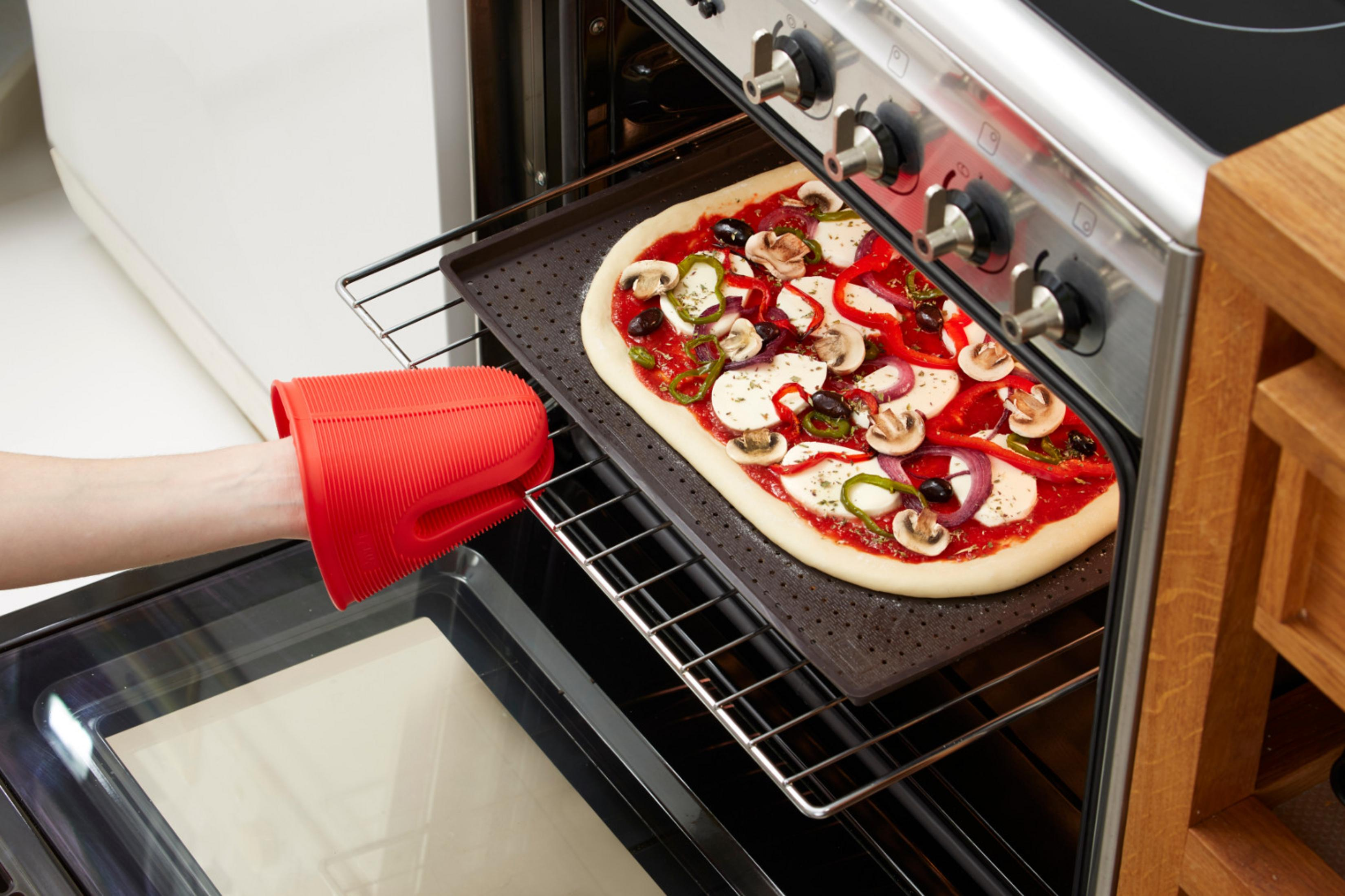 Eine Pizza mit Belag wird mit einem roten Silikon-Ofenhandschuh in einen Ofen geschoben.