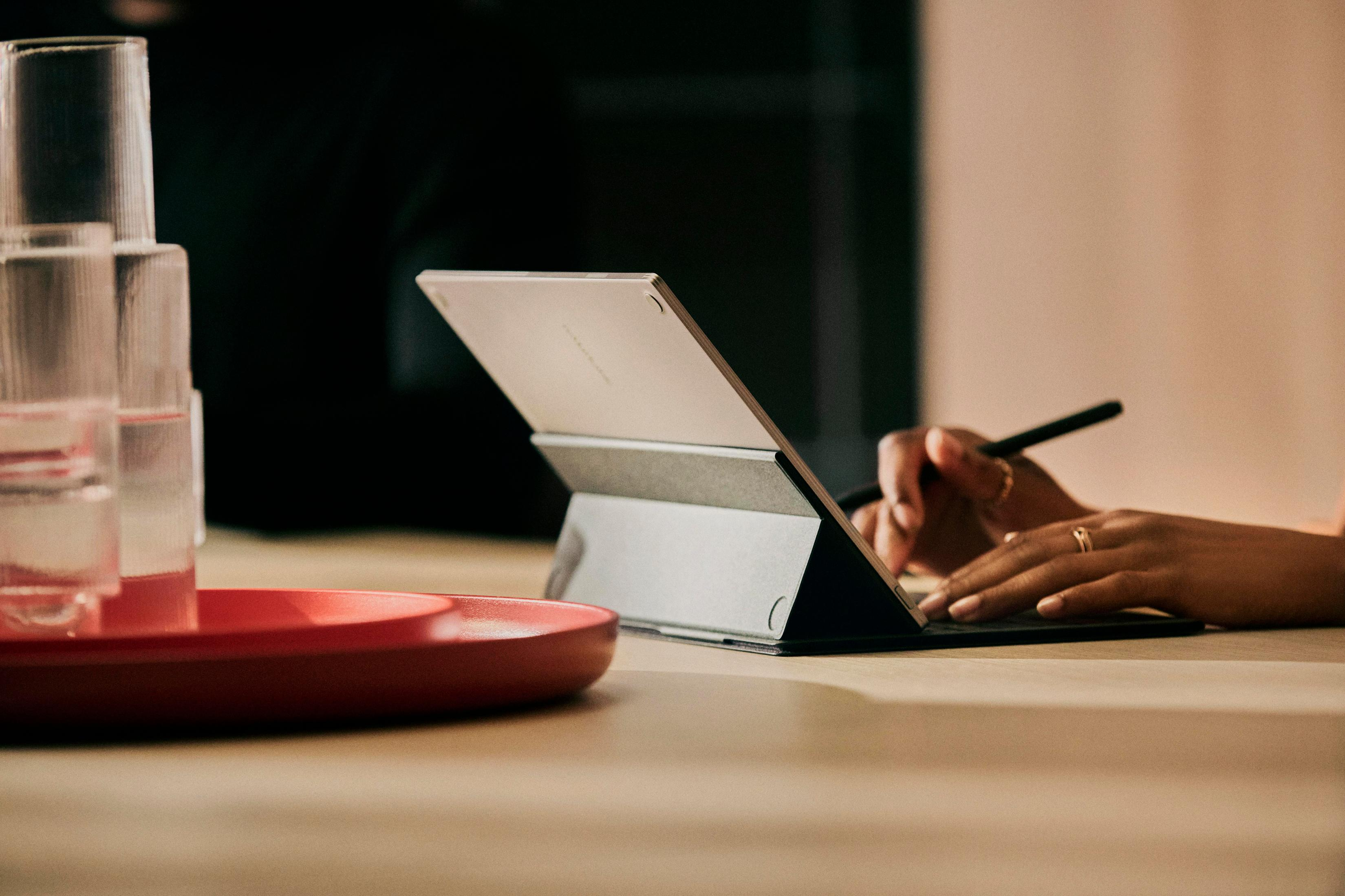 Bir kişi bir tablet üzerine bir kalemle yazıyor. Ön planda kırmızı bir tepsi ve bardaklar.