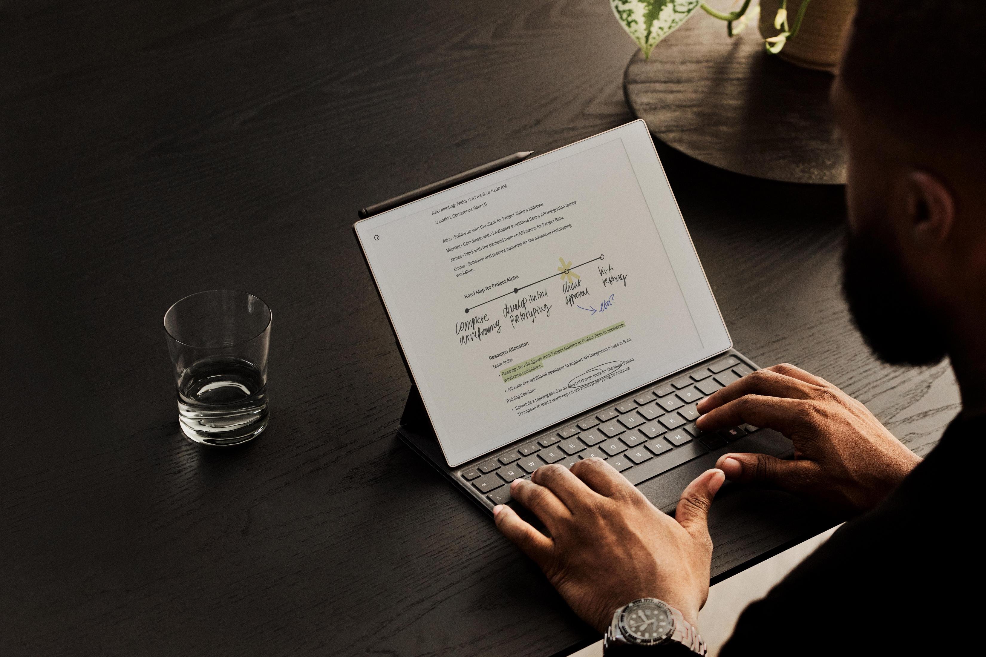 Bir kişi bir tablet klavyesinde yazıyor. Karanlık bir masada, ekranda bir bardak ve notlar.