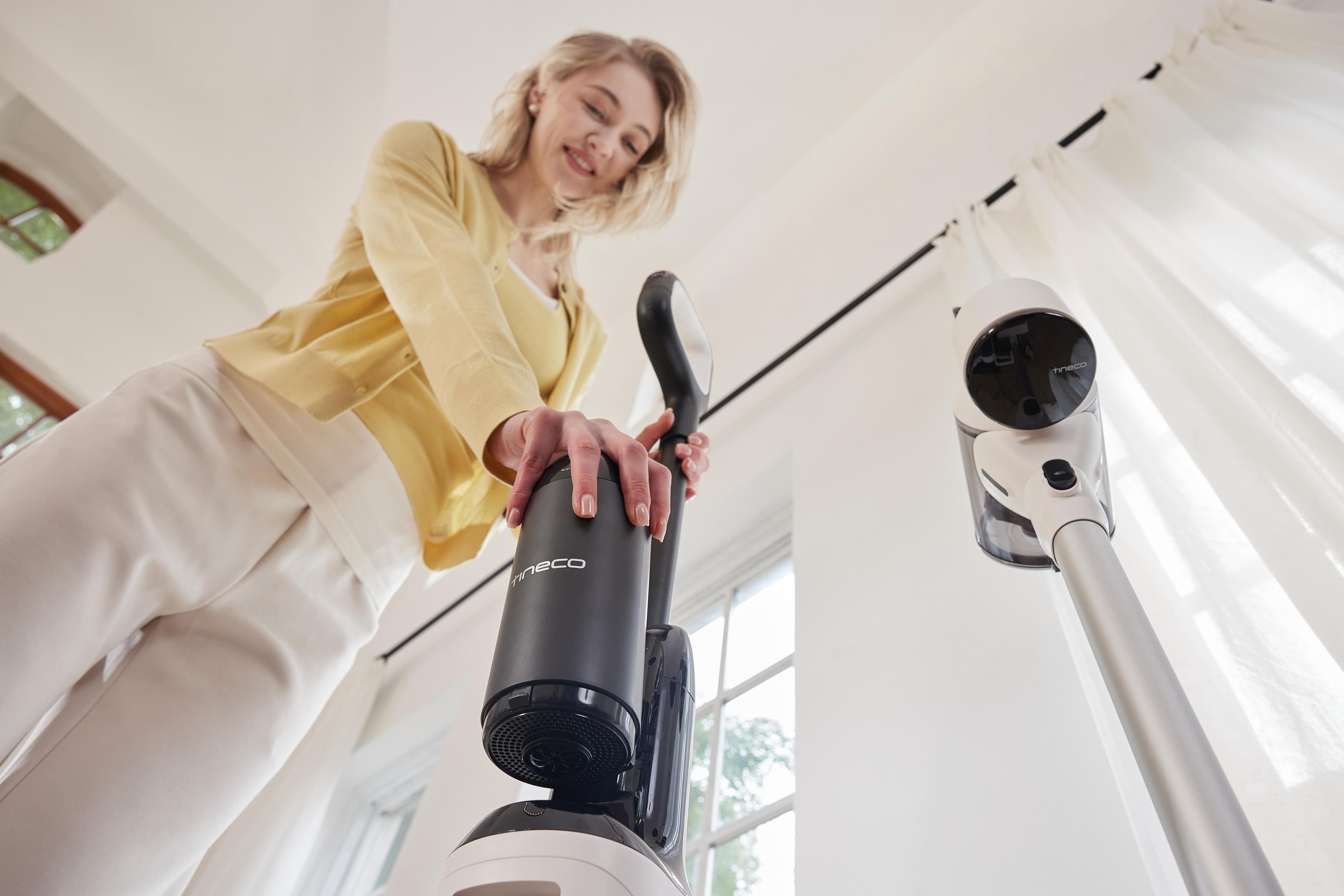 Kobieta w żółtym trzyma odkurzacz z niskiego kąta. Pokój jest biały z oknem.