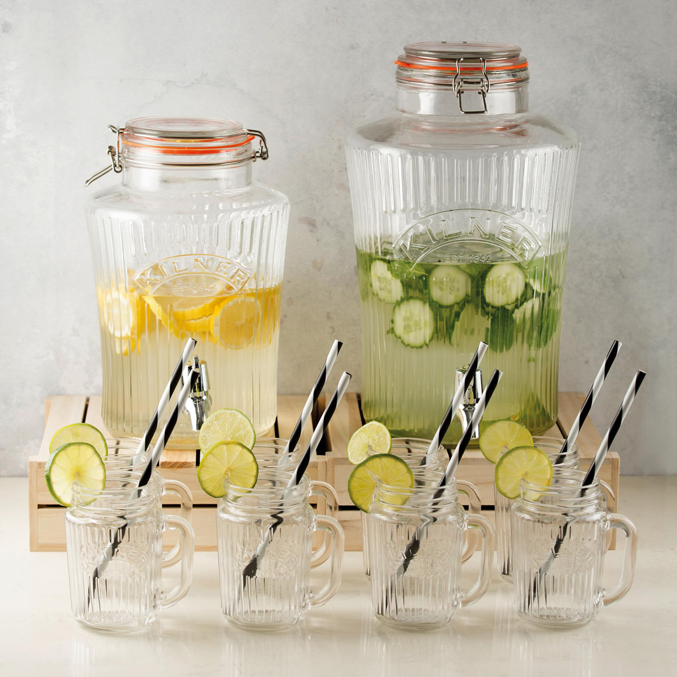 Zwei große Getränkespender aus Glas mit Ausgüssen, Zitronen, Gurken, Strohhalmen und kleinen Glasbechern.