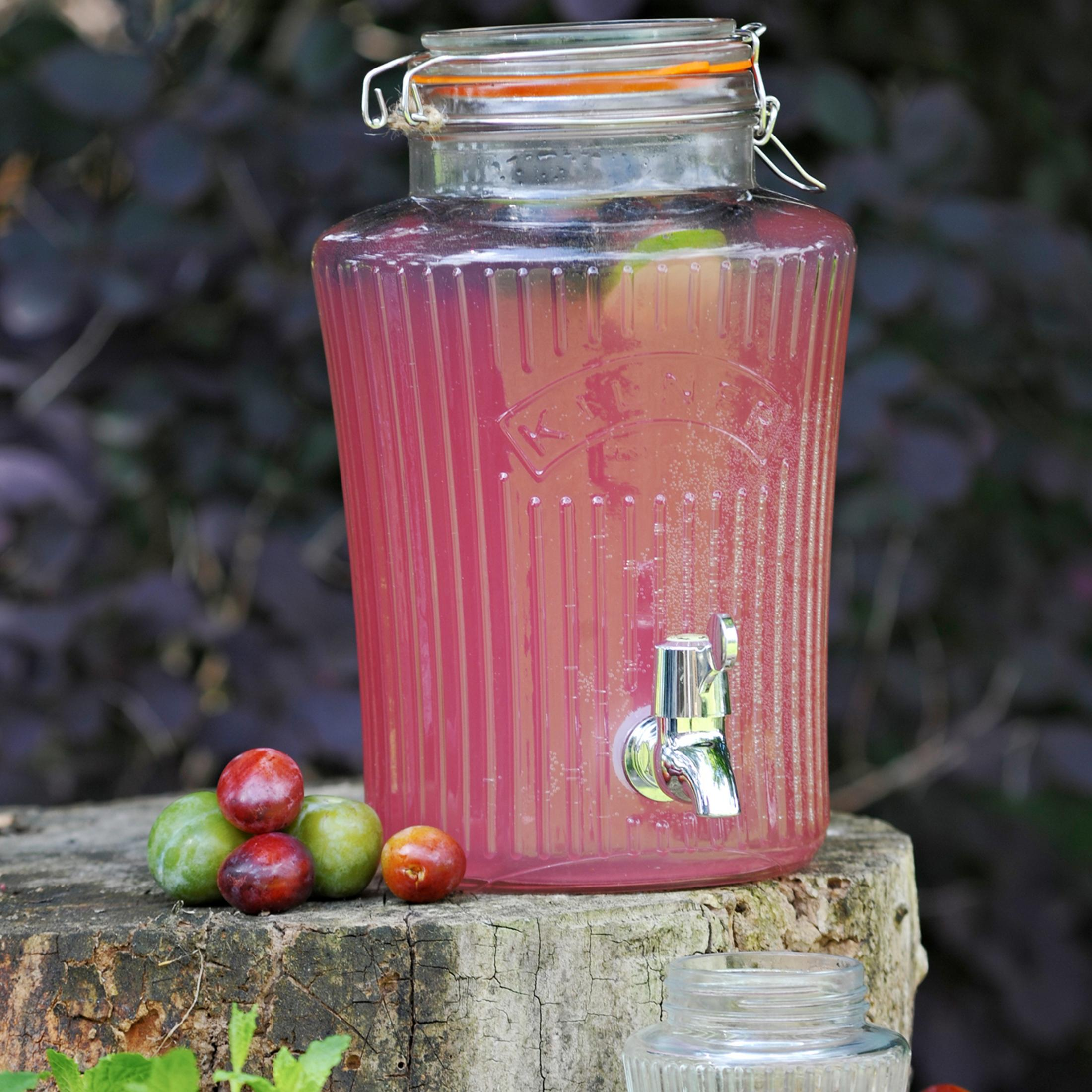 Ein Getränkespender aus Glas, gefüllt mit einem rosa Getränk, der auf einem Baumstumpf mit Pflaumen steht.