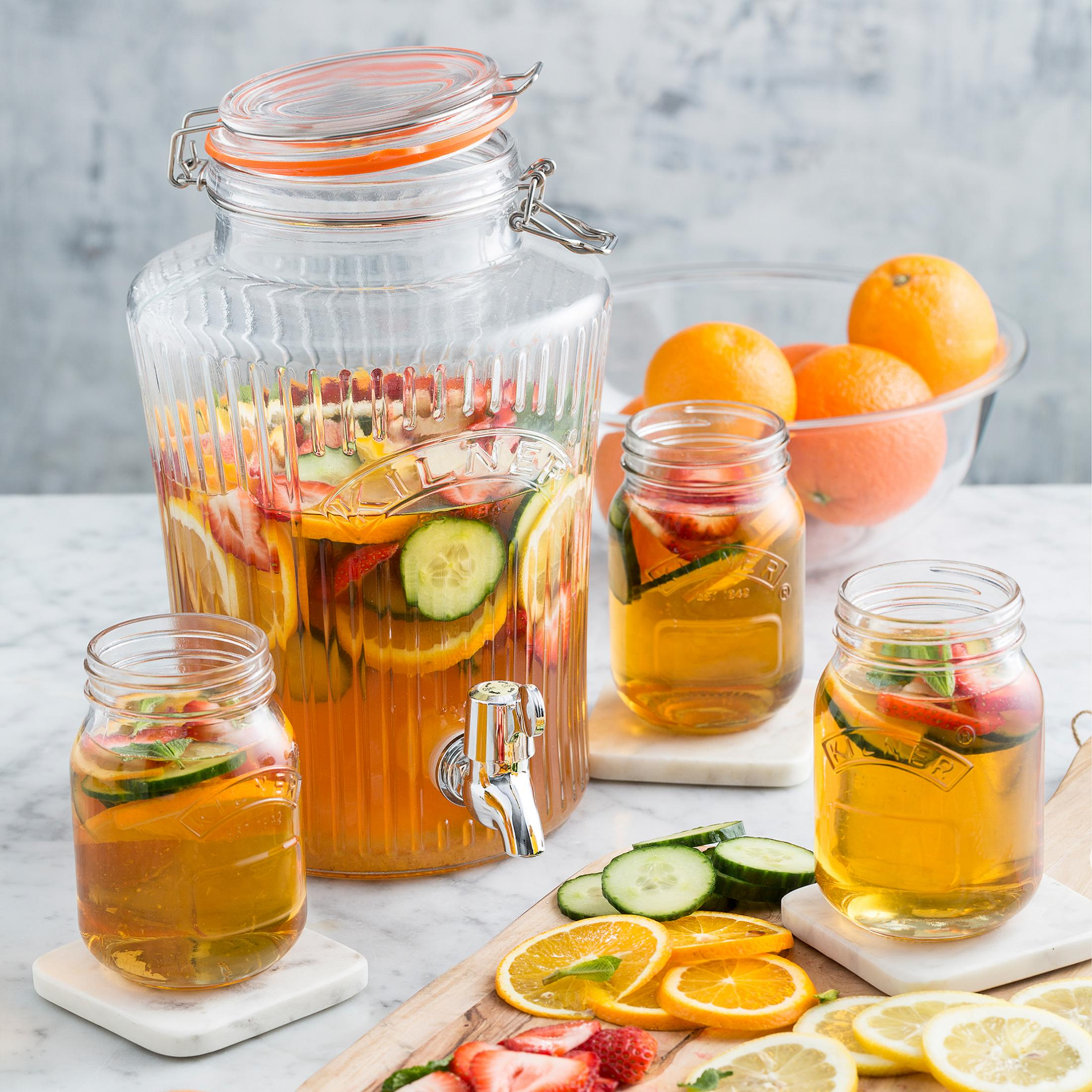 Getränkespender aus Glas mit Früchten, plus 3 gefüllte Becher, Orangen und geschnittene Früchte.
