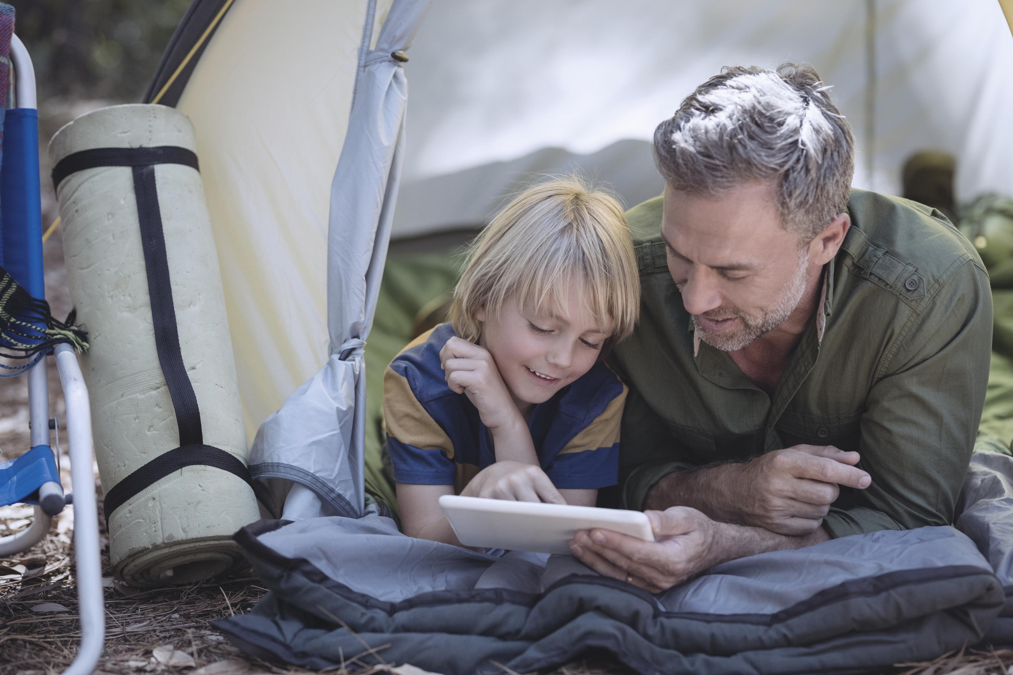 Vater und Sohn im Zelt schauen auf ein Tablet. Grünes und gelbes Zelt. Hintergrund Wald.