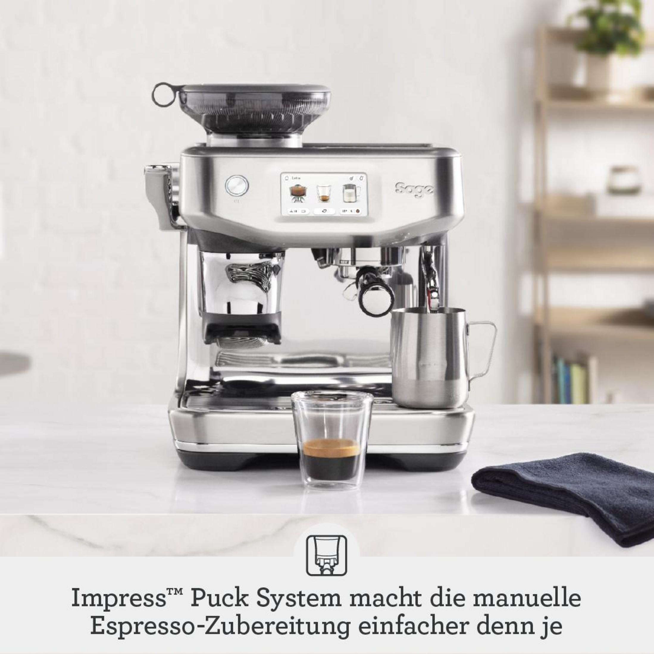 Una macchina da caffè Sage argentata si erge su un tavolo bianco con un piccolo bicchiere di espresso e un asciugamano blu scuro.