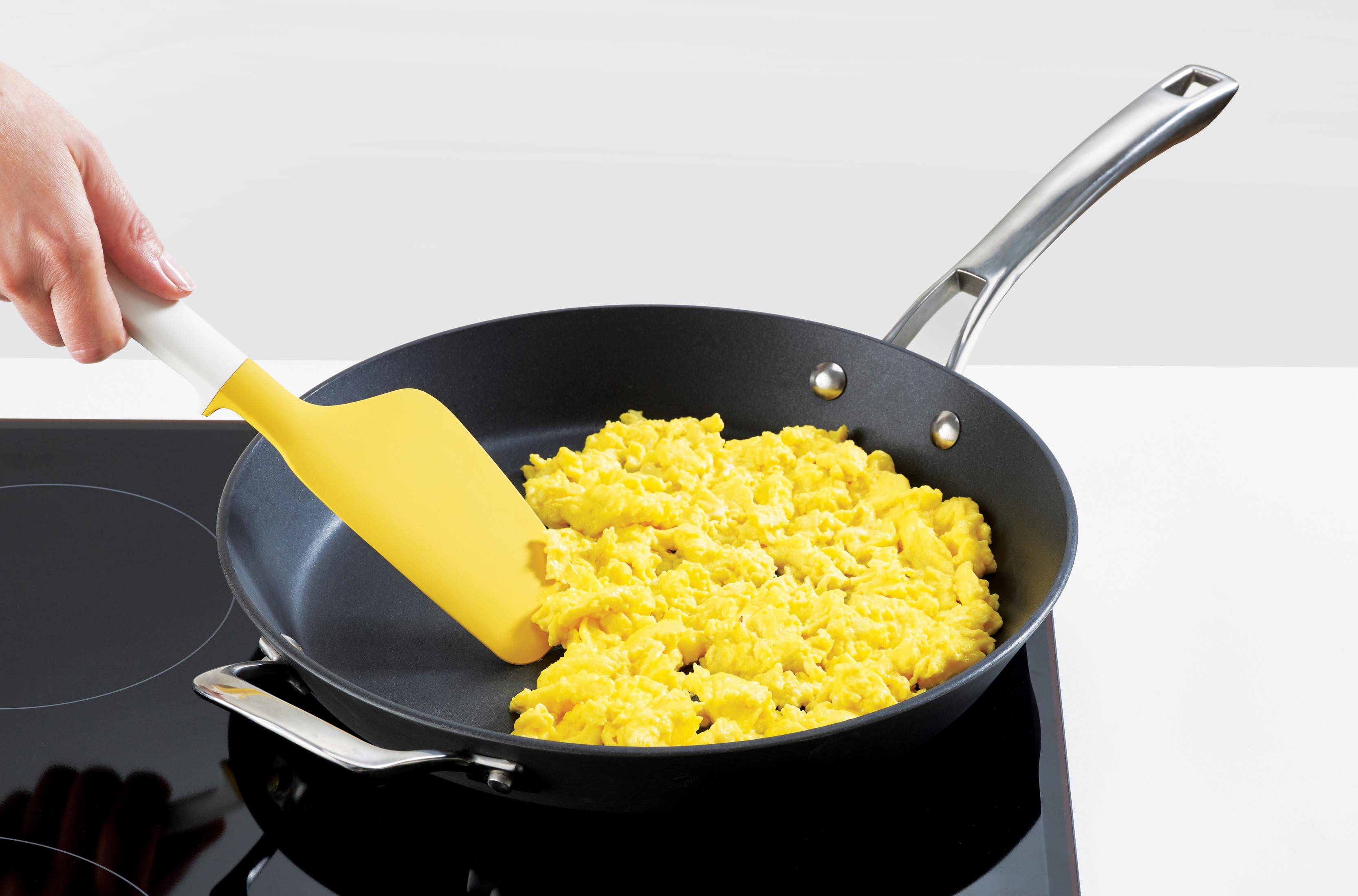 Sarı spatula, ocağın üzerinde siyah bir tavada çırpılmış yumurtaları karıştırıyor. Bir el spatulayı tutuyor.