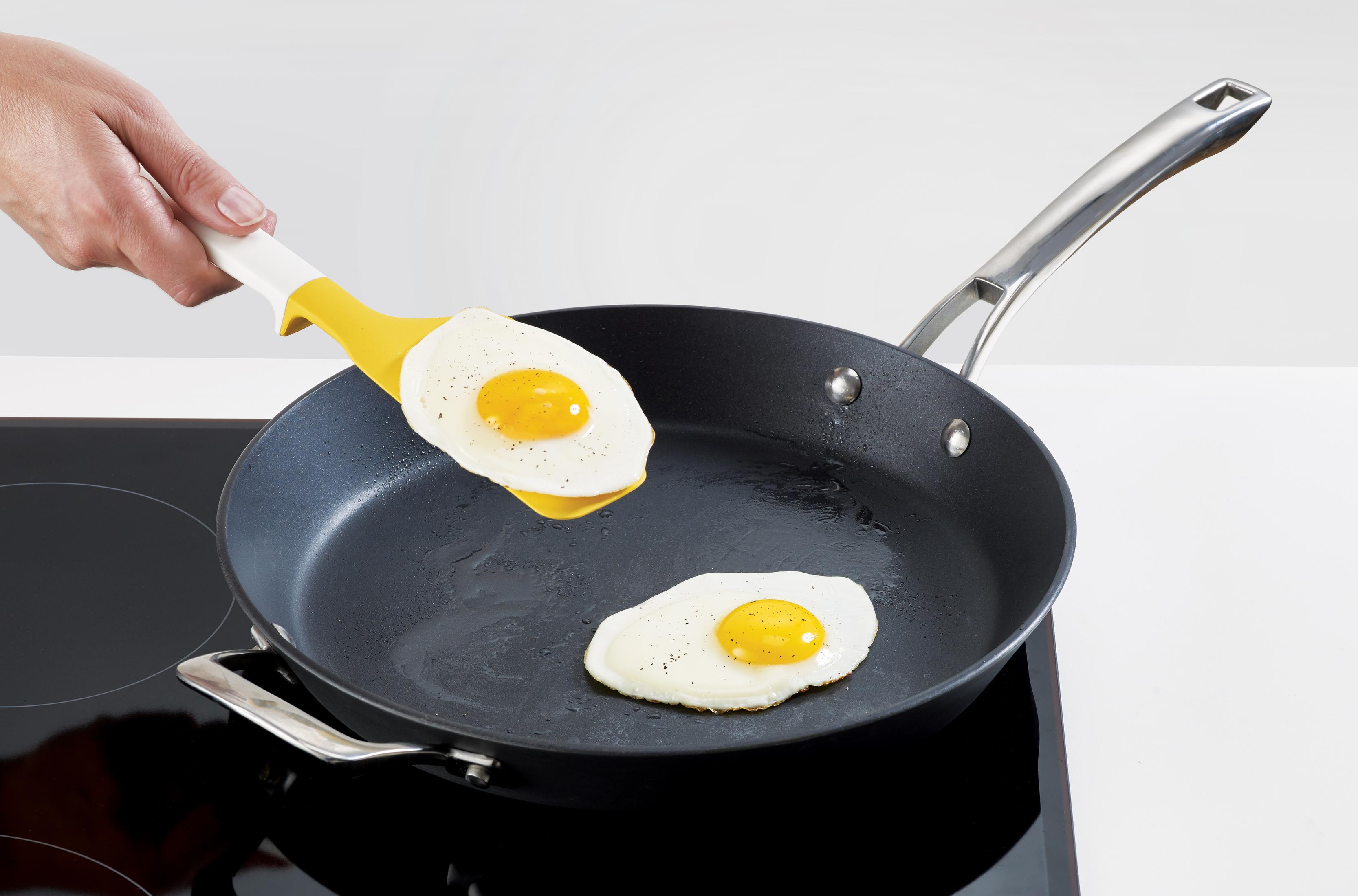 Sarı spatula, ocaktaki siyah bir tavadan iki kızarmış yumurtayı çıkarıyor. Yumurtalar beyaz bir arka planda.