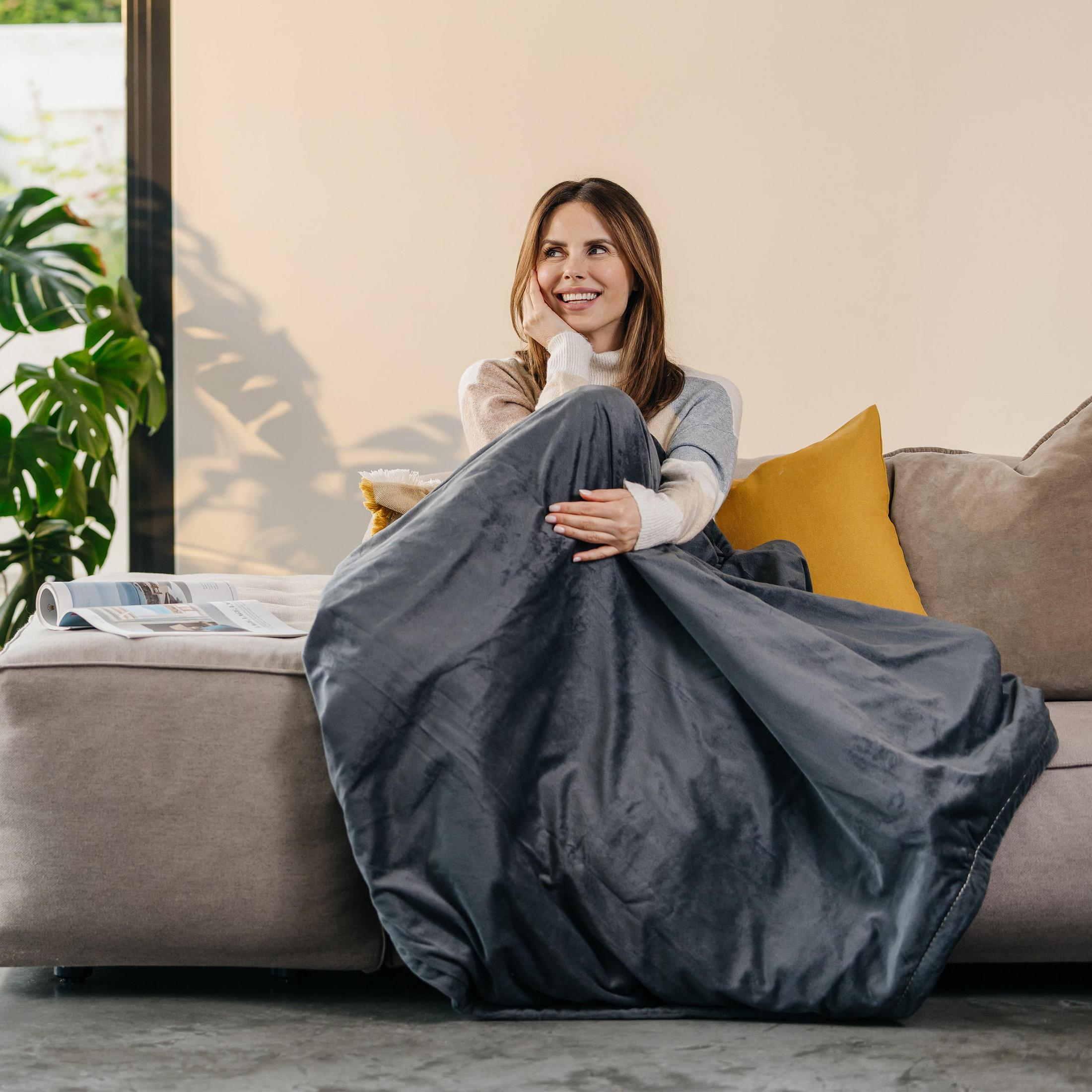 Eine Frau lächelt, eingewickelt in eine graue Heizdecke auf einer grauen Couch im Innenbereich.
