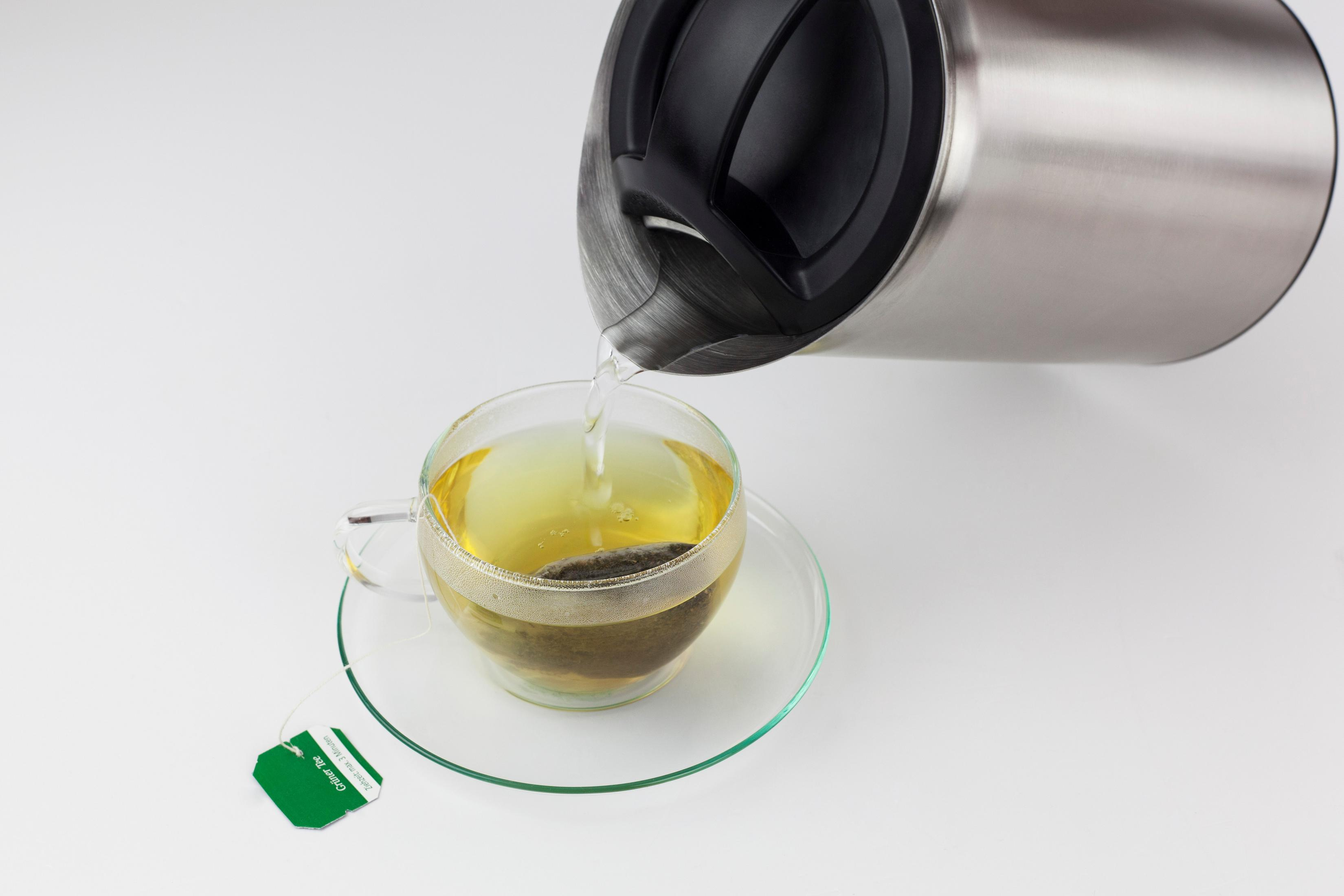 Ein silberner Wasserkocher gießt heißes Wasser in eine durchsichtige Glastasse mit einem Teebeutel auf einer Untertasse.