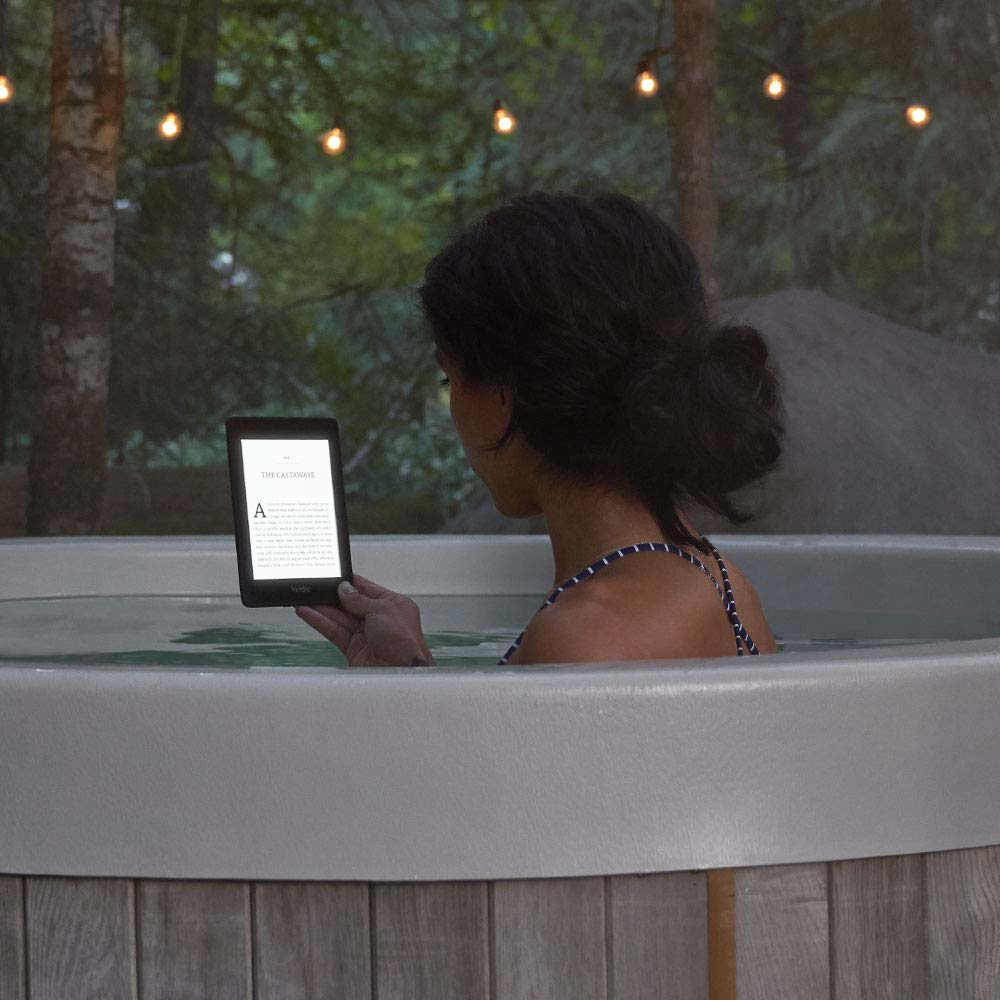 Frau liest einen Kindle in einem Whirlpool, umgeben von Bäumen und Lichterketten in der Abenddämmerung.